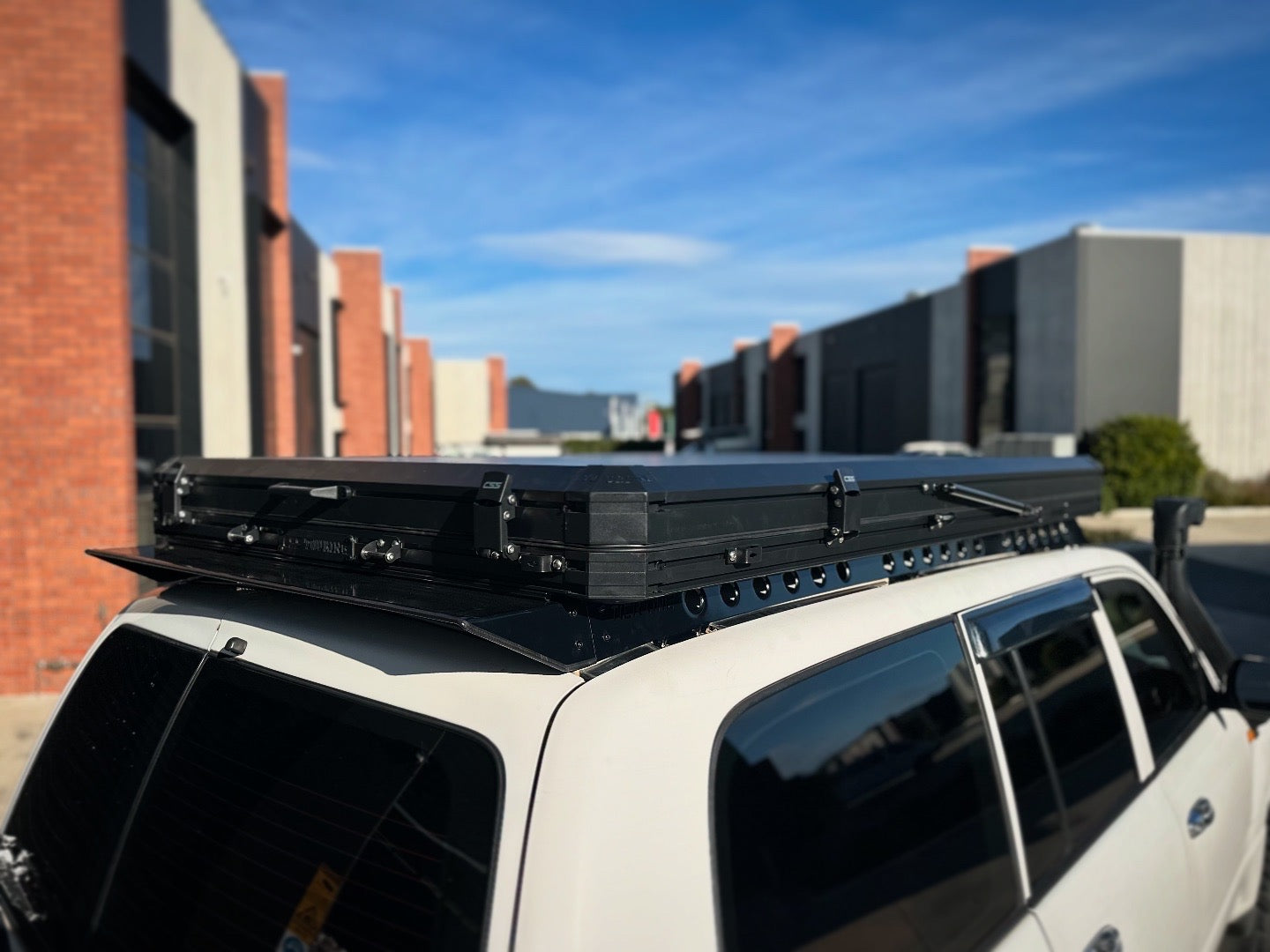 This is an image of a white 4x4 fitted with a black Tier One Metalworks Roof Rack & Rails under a closed rooftop tent in an industrial area