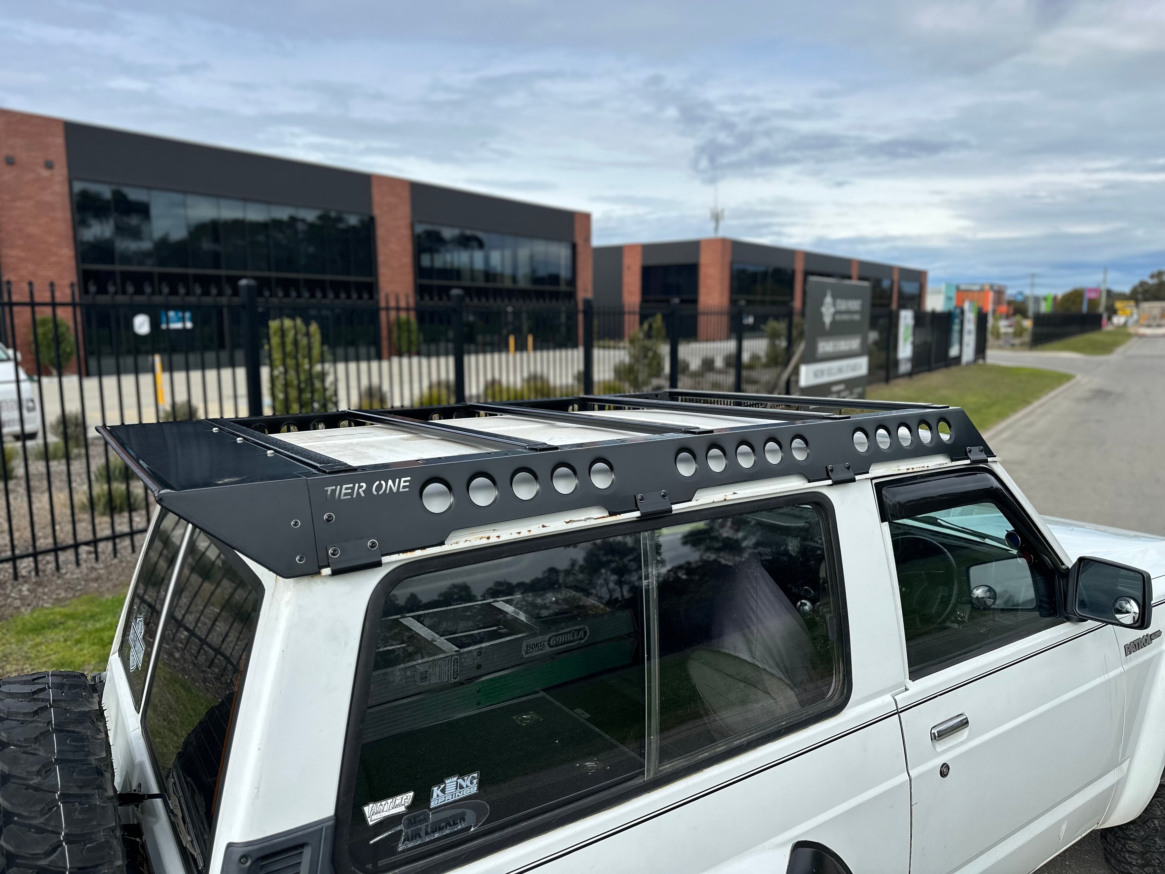 This is an image of a white Nissan Patrol fitted with a black Tier One Metalworks Roof Rack on an urban street.