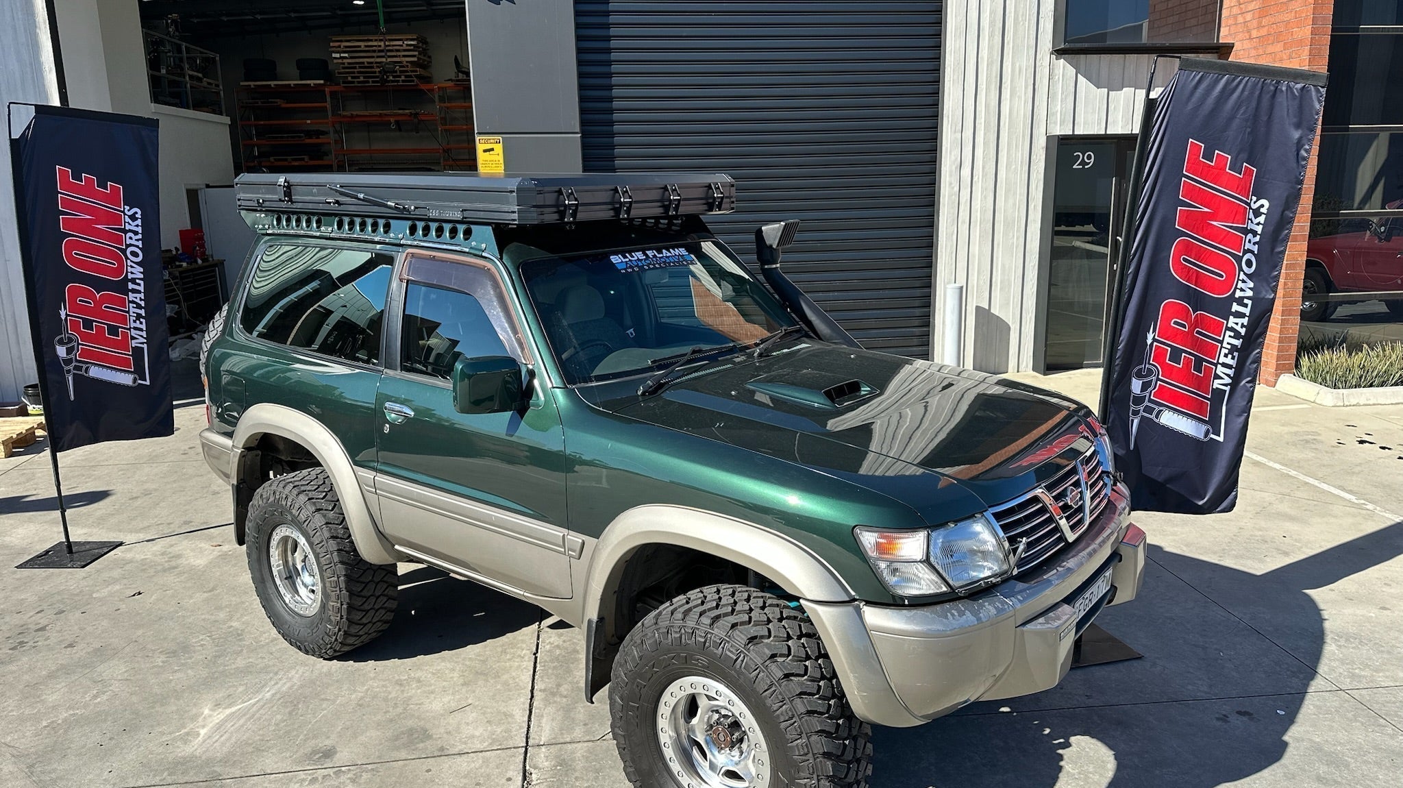 This is an image of a green and beige Nissan Patrol with Tier One Metalworks roof racks parked outside a workshop