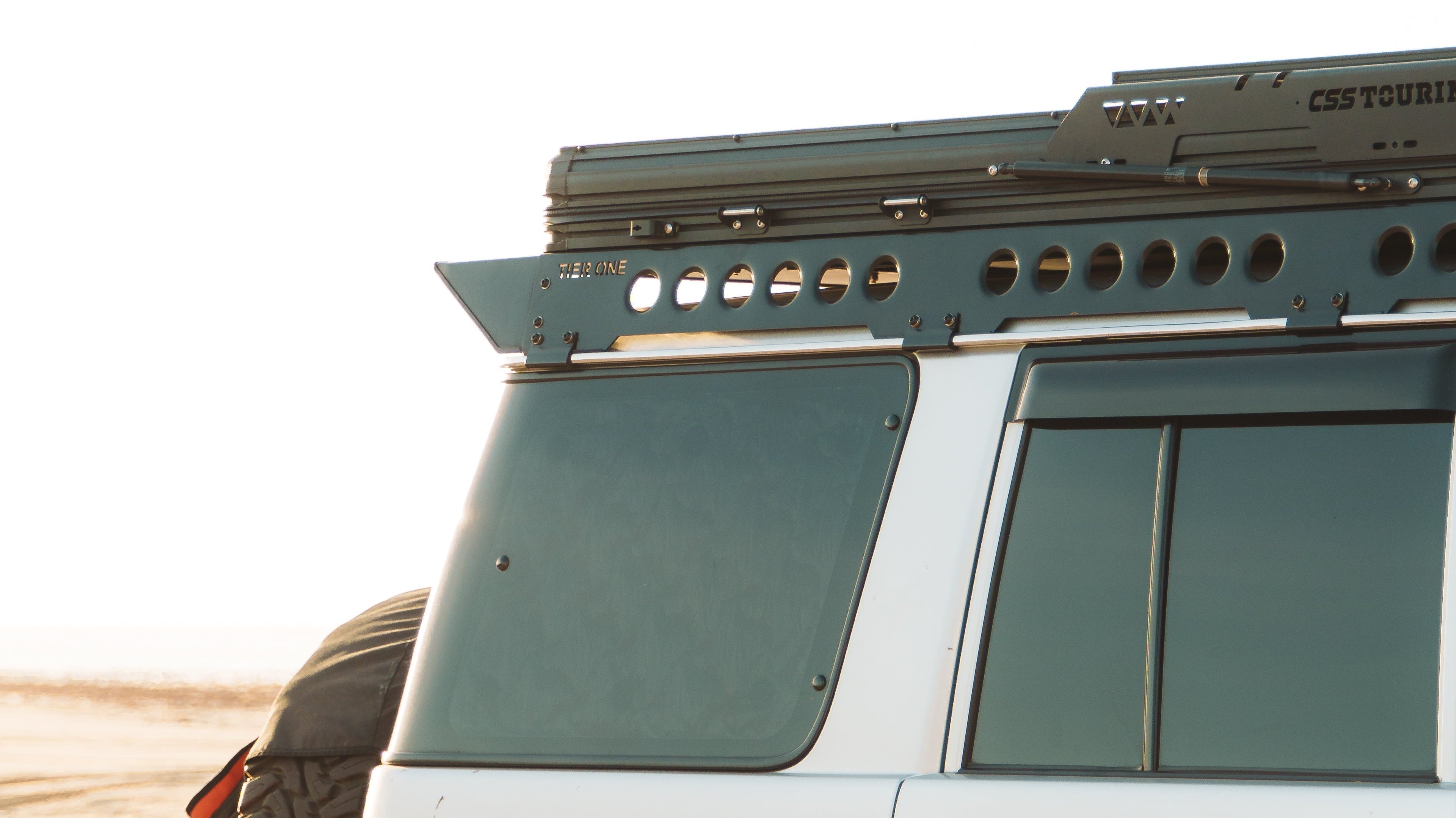 This is an image of a white 4x4 vehicle equipped with Tier One Metalworks Roof Racks & Rails and a CSS Touring rooftop tent on sandy terrain.