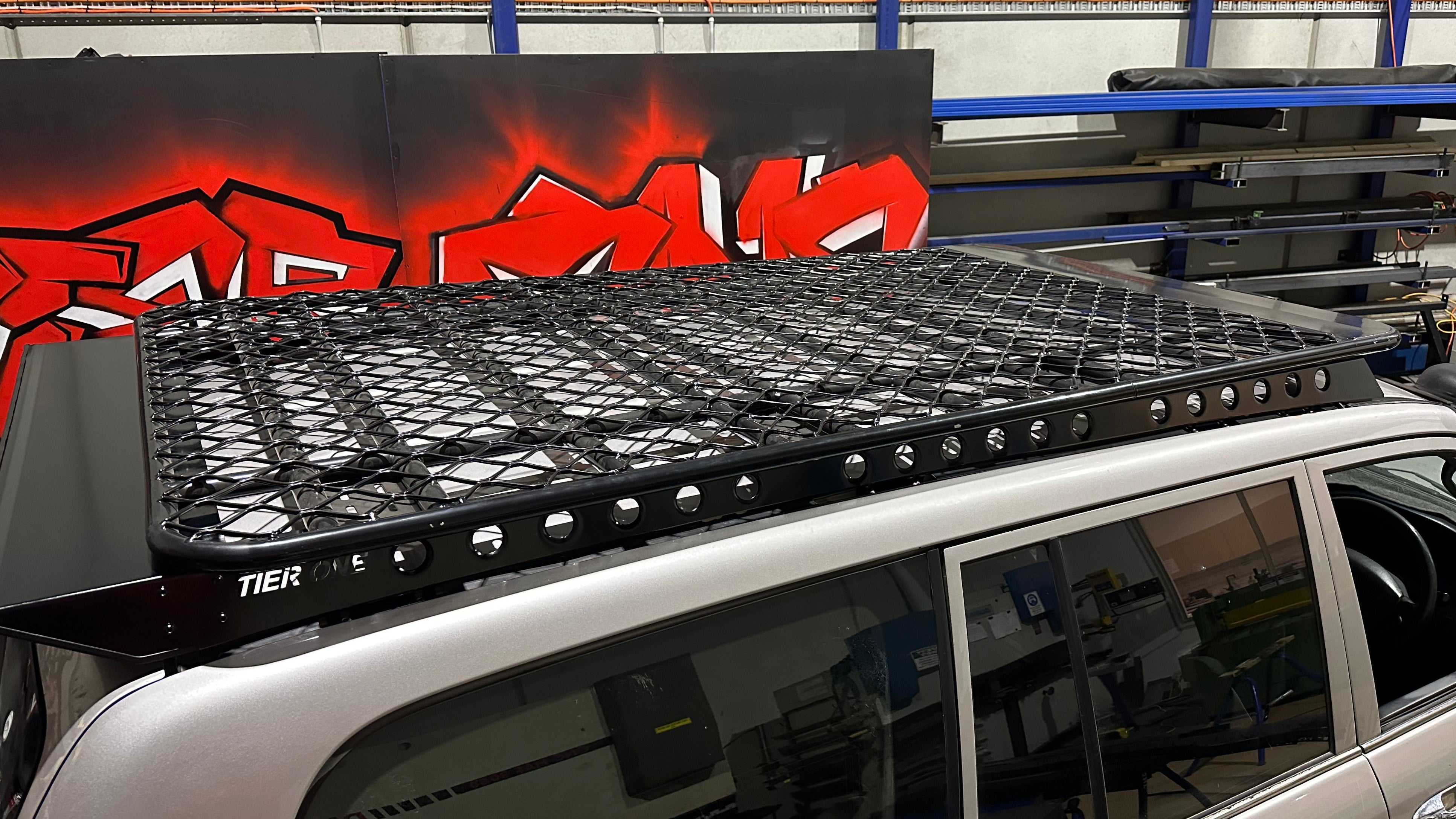 This is an image of a black Tier One Metalworks steel roof rack with mesh design mounted on a silver 4WD in a workshop with red graffiti wall