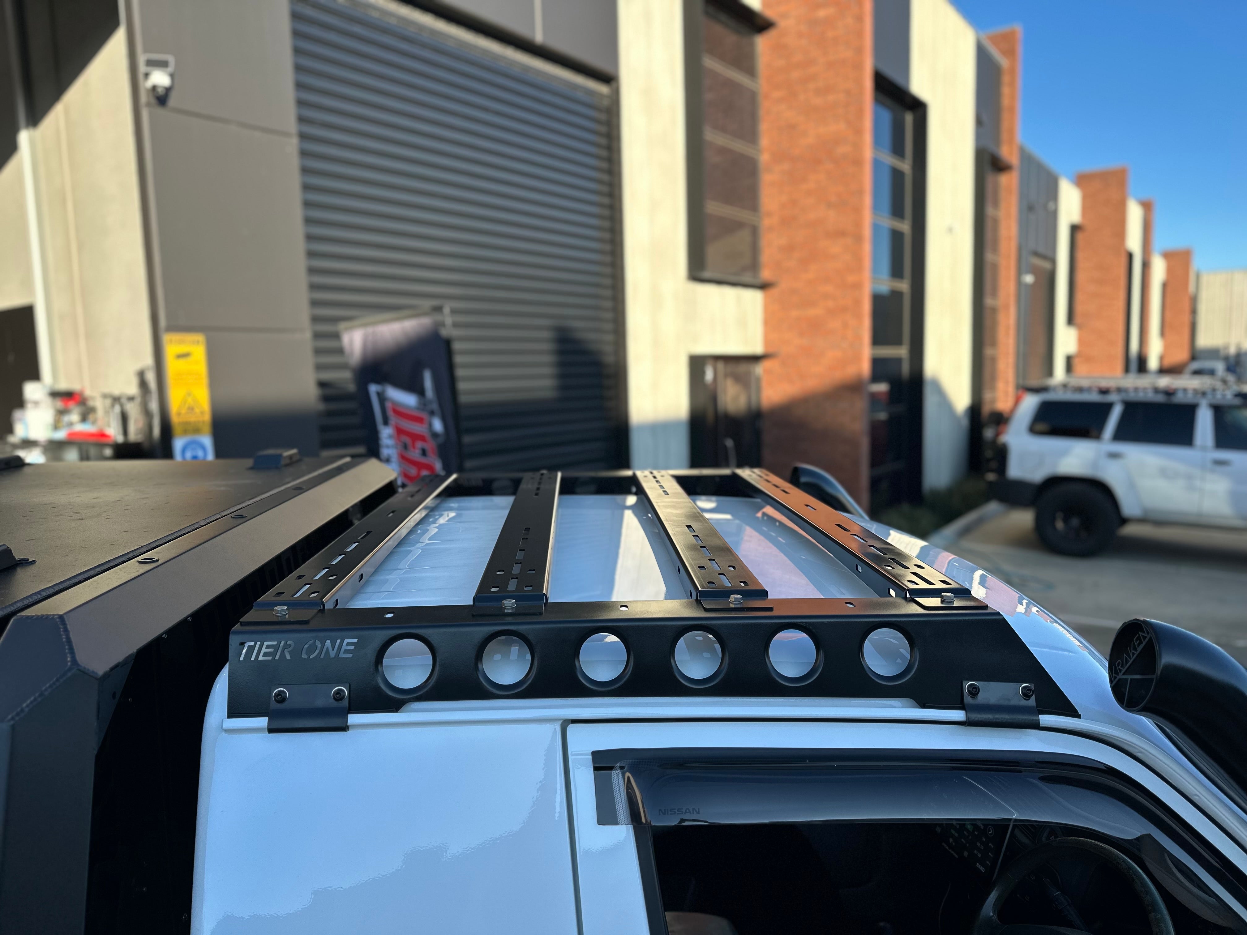 This is an image of a white 4x4 vehicle with a black Tier One Metalworks Roof Rack on the roof, parked outside an industrial building in Australia