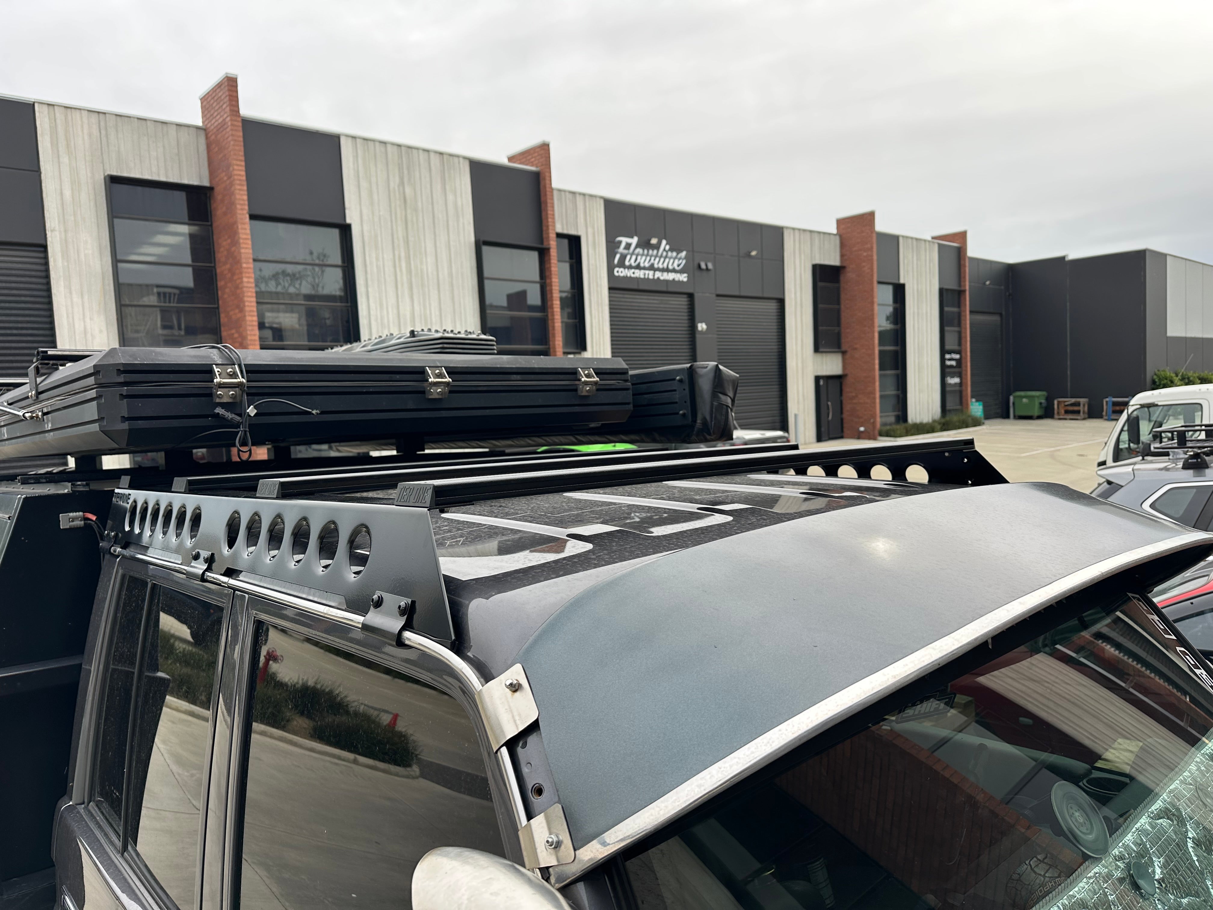 This is an image of a grey 4x4 vehicle fitted with Tier One Metalworks black metal roof racks and rails in an industrial area
