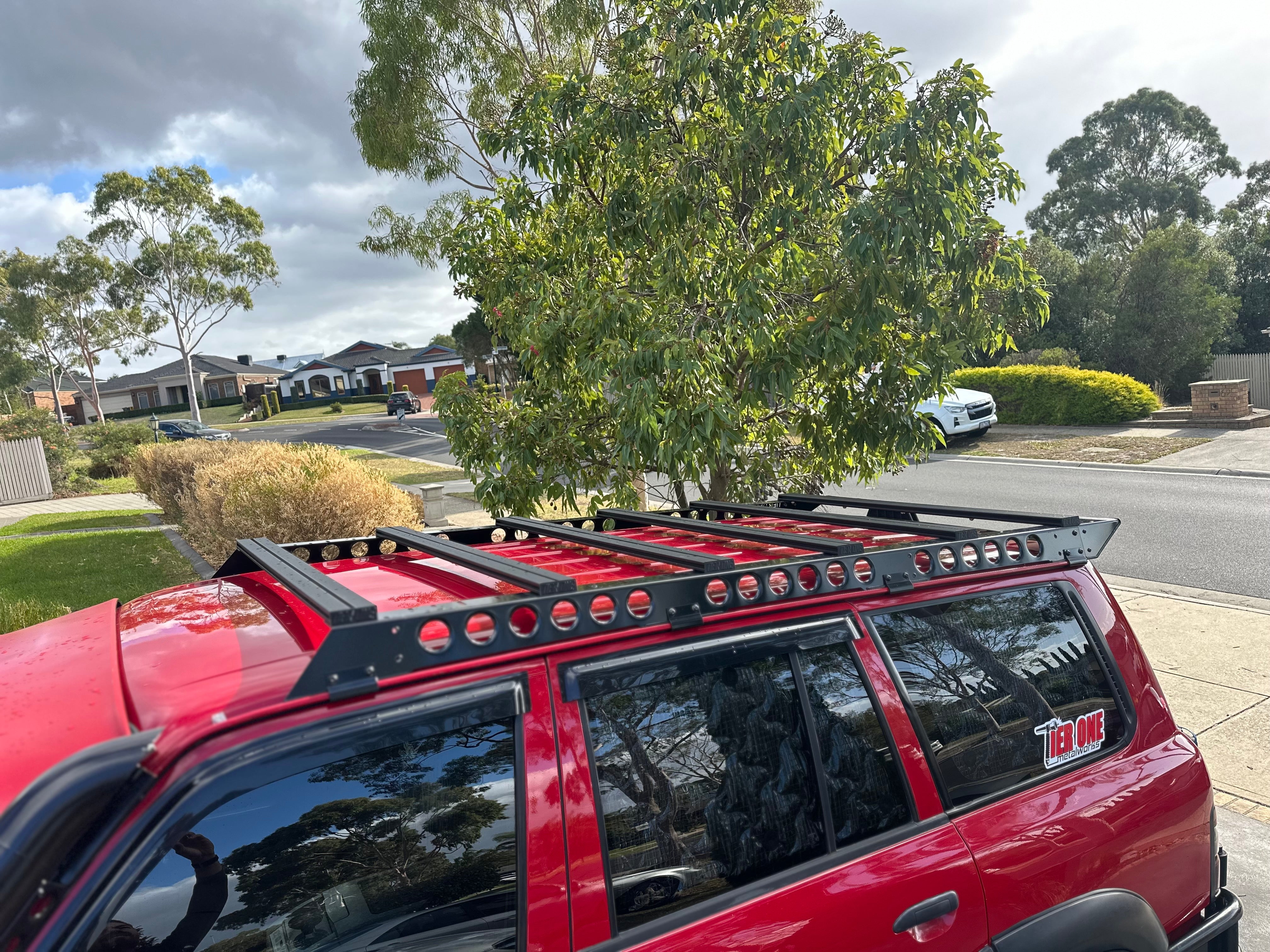 This is an image of a red 4WD with a black Tier One Metalworks roof rack installed outdoors on a cloudy day