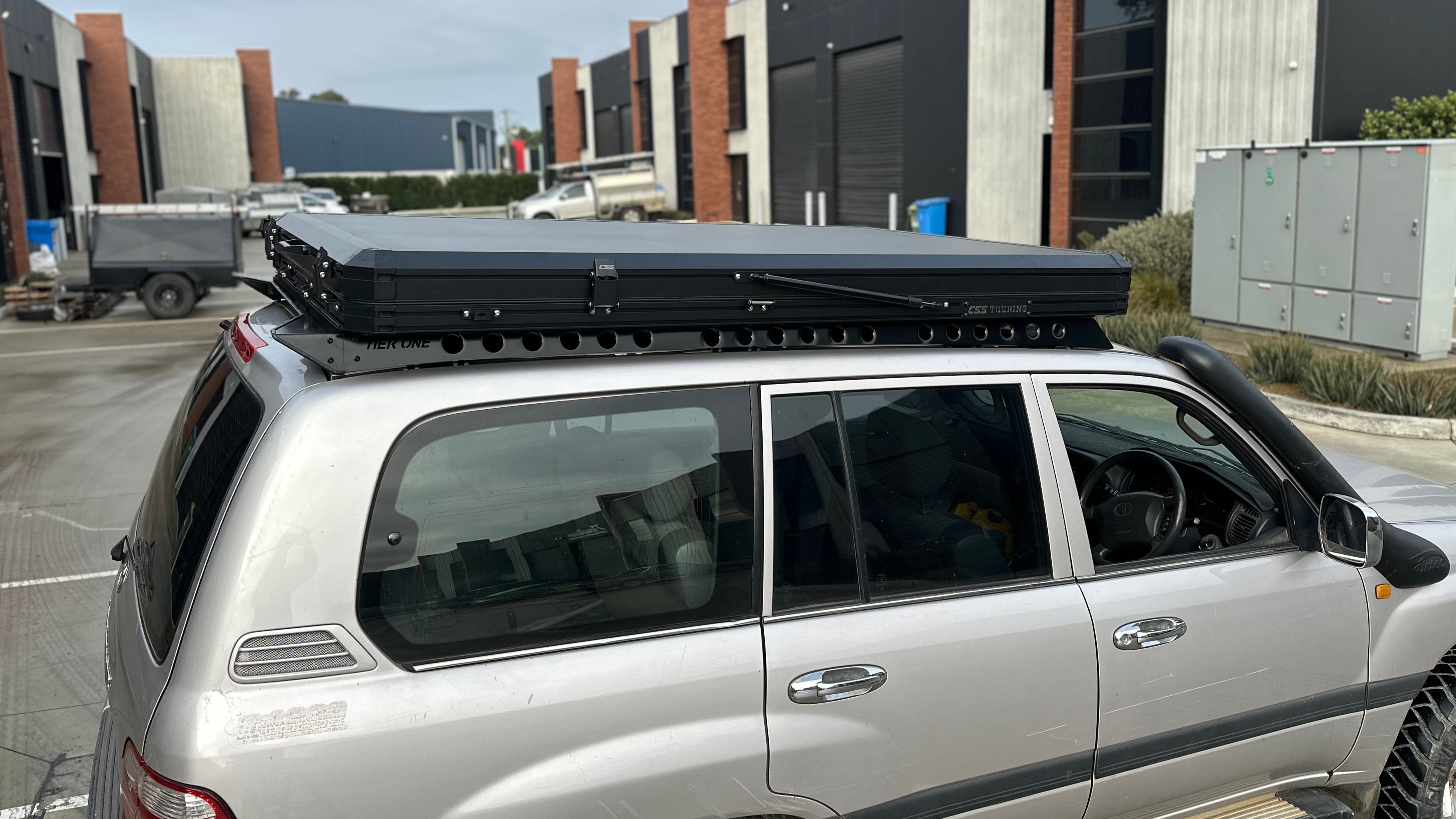 This is an image of a silver Toyota Land Cruiser fitted with Tier One Metalworks black roof racks and a CSS Touring rooftop tent in an industrial setting
