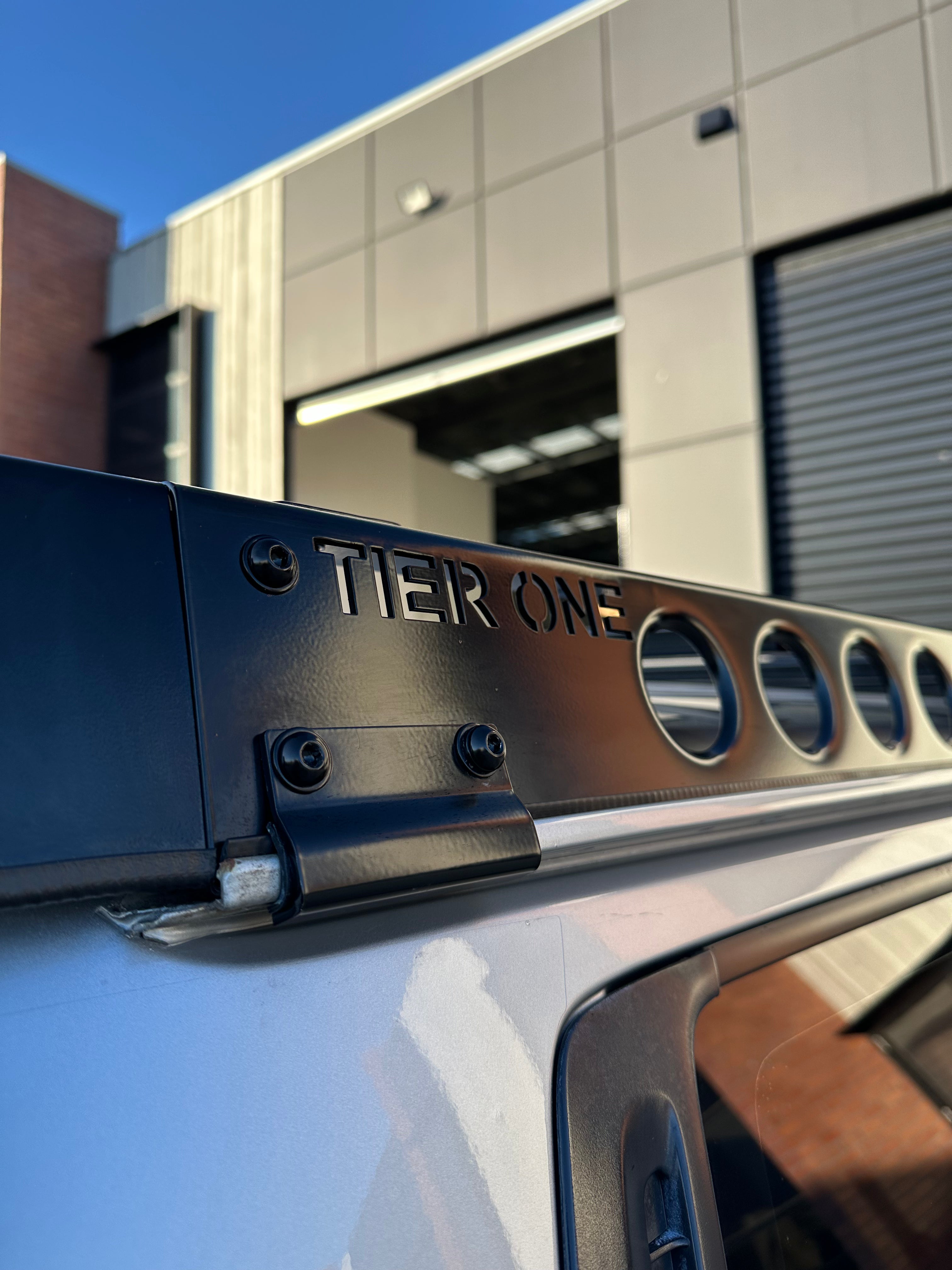 This is an image of a black Tier One Metalworks roof rack mounted on a silver 4WD vehicle in an industrial setting