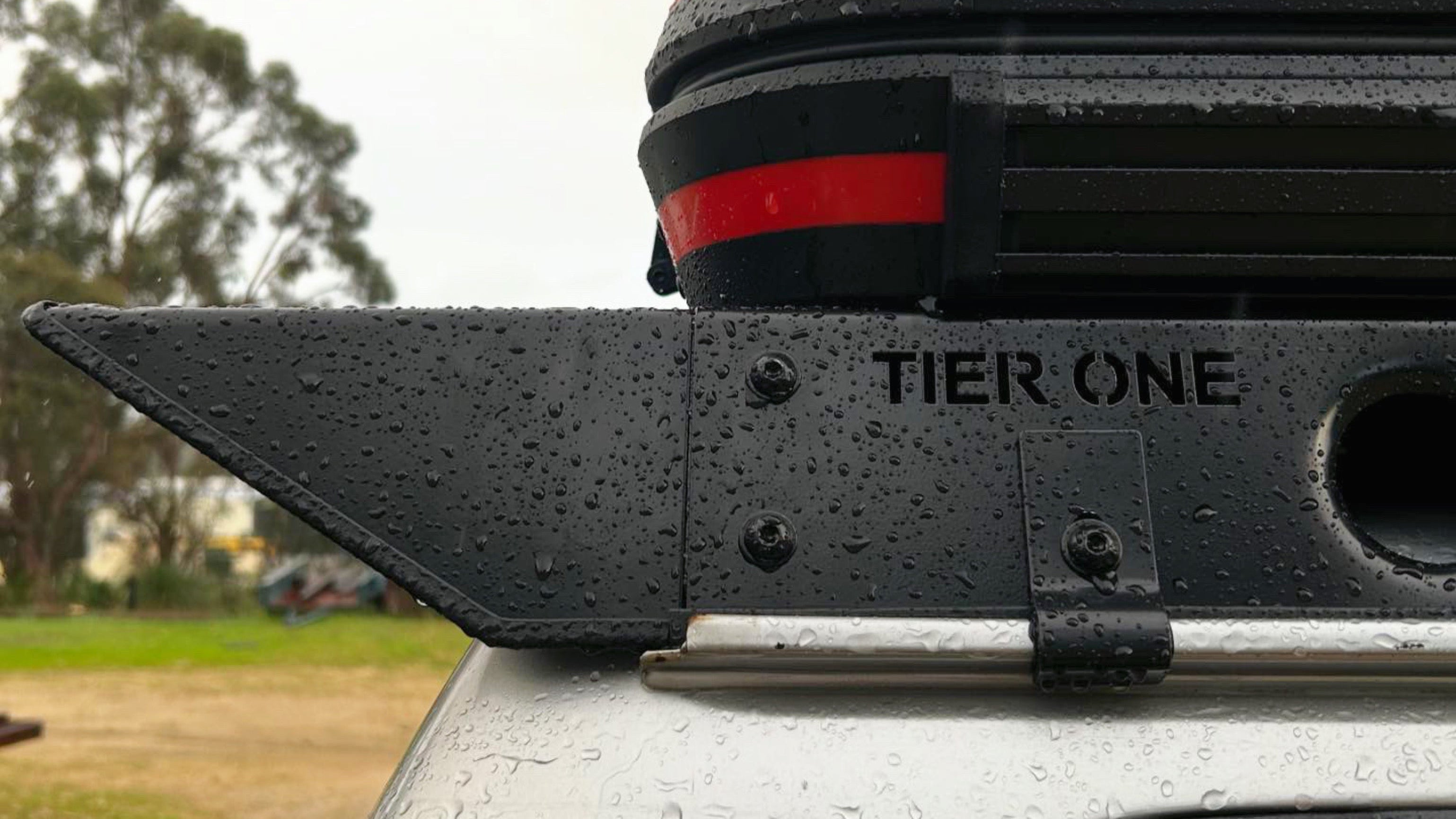 This is an image of a black Tier One Metalworks roof rack mounted on a silver 4WD vehicle, wet with rain droplets
