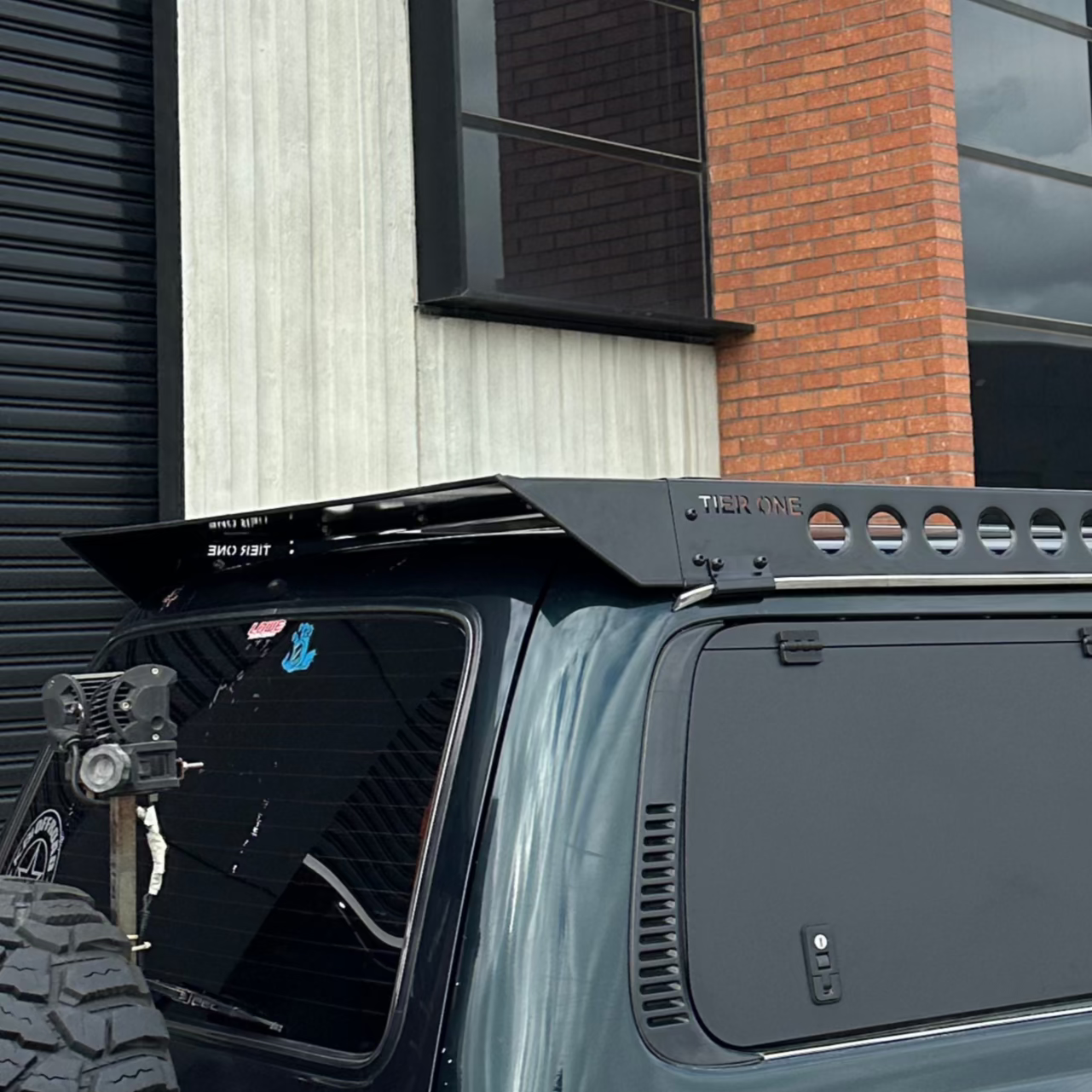 This is an image of a black Tier One Metalworks roof rack and rails mounted on a dark green 4x4 vehicle against an industrial building backdrop