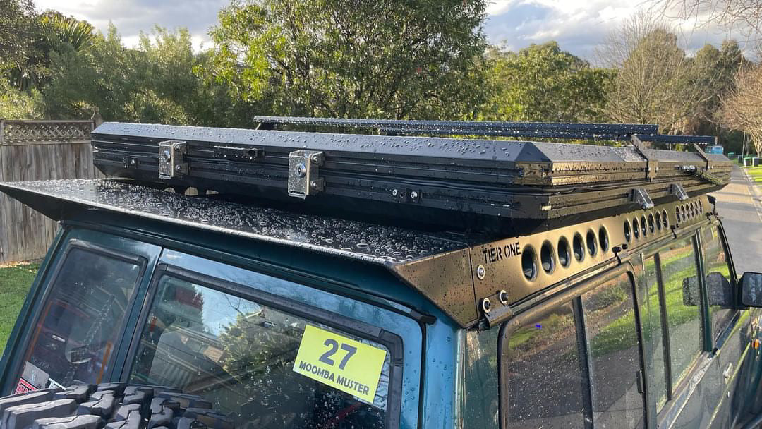 This is an image of a blue 4x4 fitted with a black Tier One Metalworks Roof Rack and Rails feature with water droplets on a rainy day
