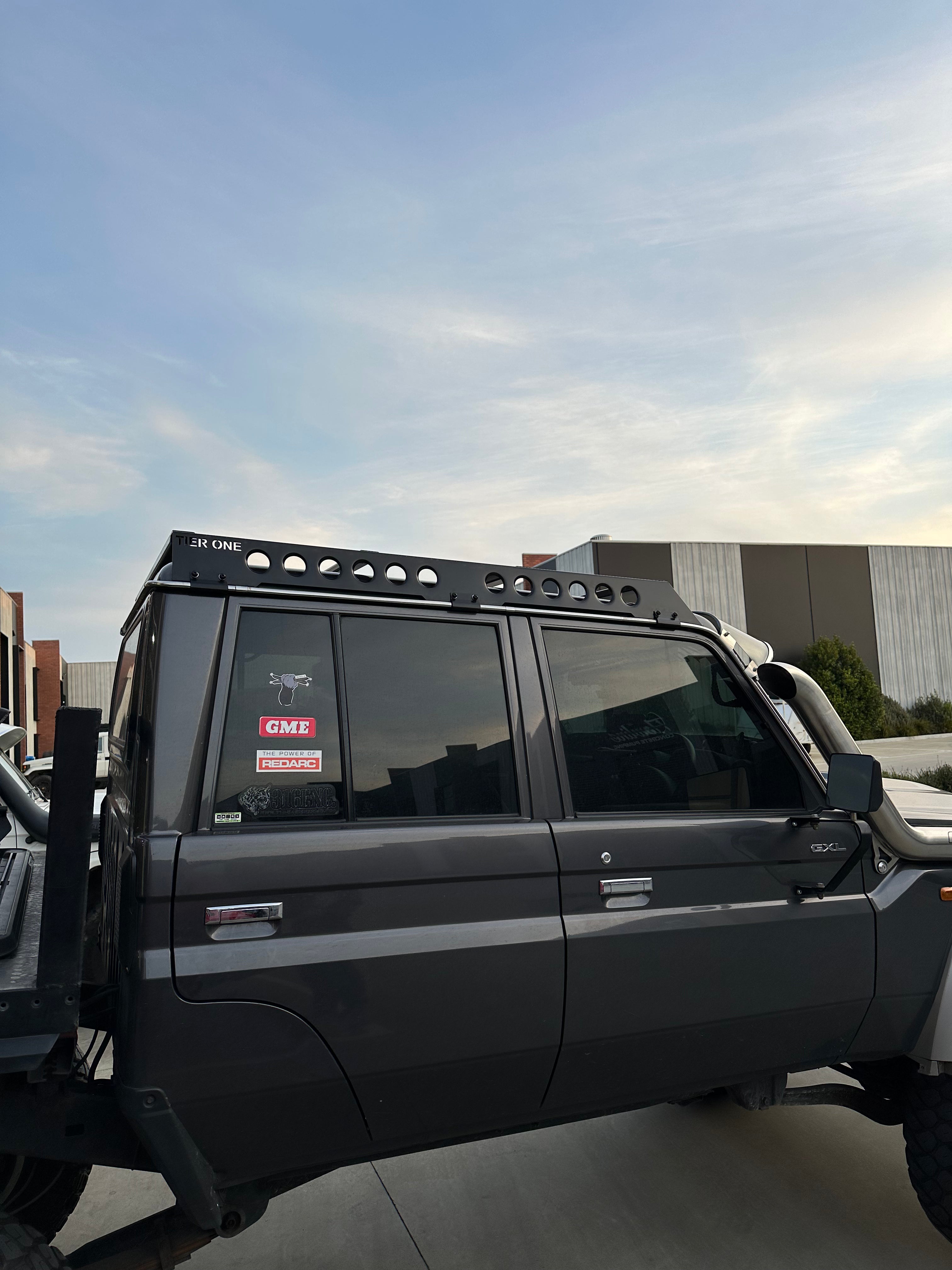 This is an image of a dark grey 4x4 with Tier One Metalworks roof rack mounted, showing side view with stickers on rear window