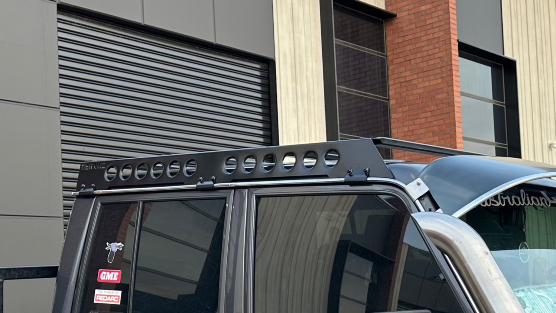 This is an image of a black roof rack by Tier One Metalworks mounted on a 4x4 vehicle with stickers on the rear window