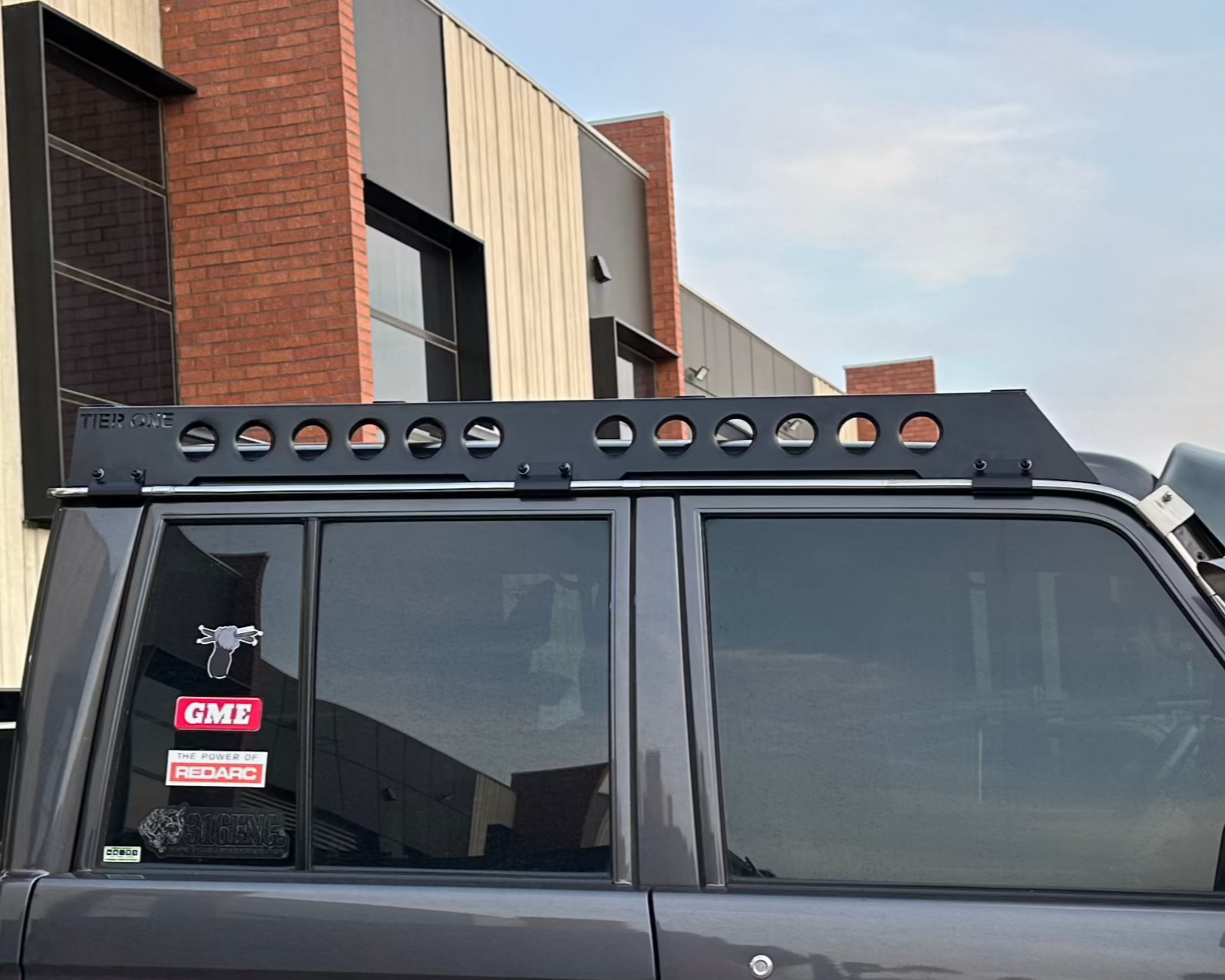 This is an image of a dark grey 4x4 vehicle fitted with a black Tier One Metalworks roof rack and rails, parked by a modern industrial building.