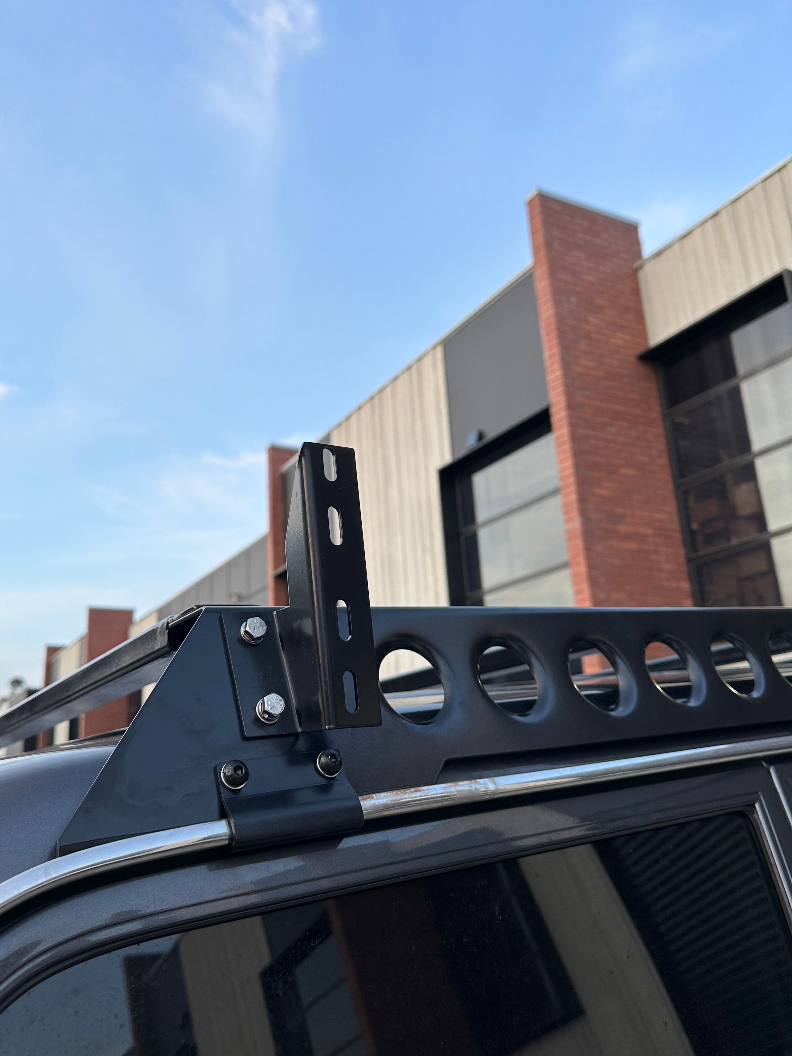 This is an image of a black Tier One Metalworks 4WD roof rack mounted on a vehicle roof with a building and blue sky background