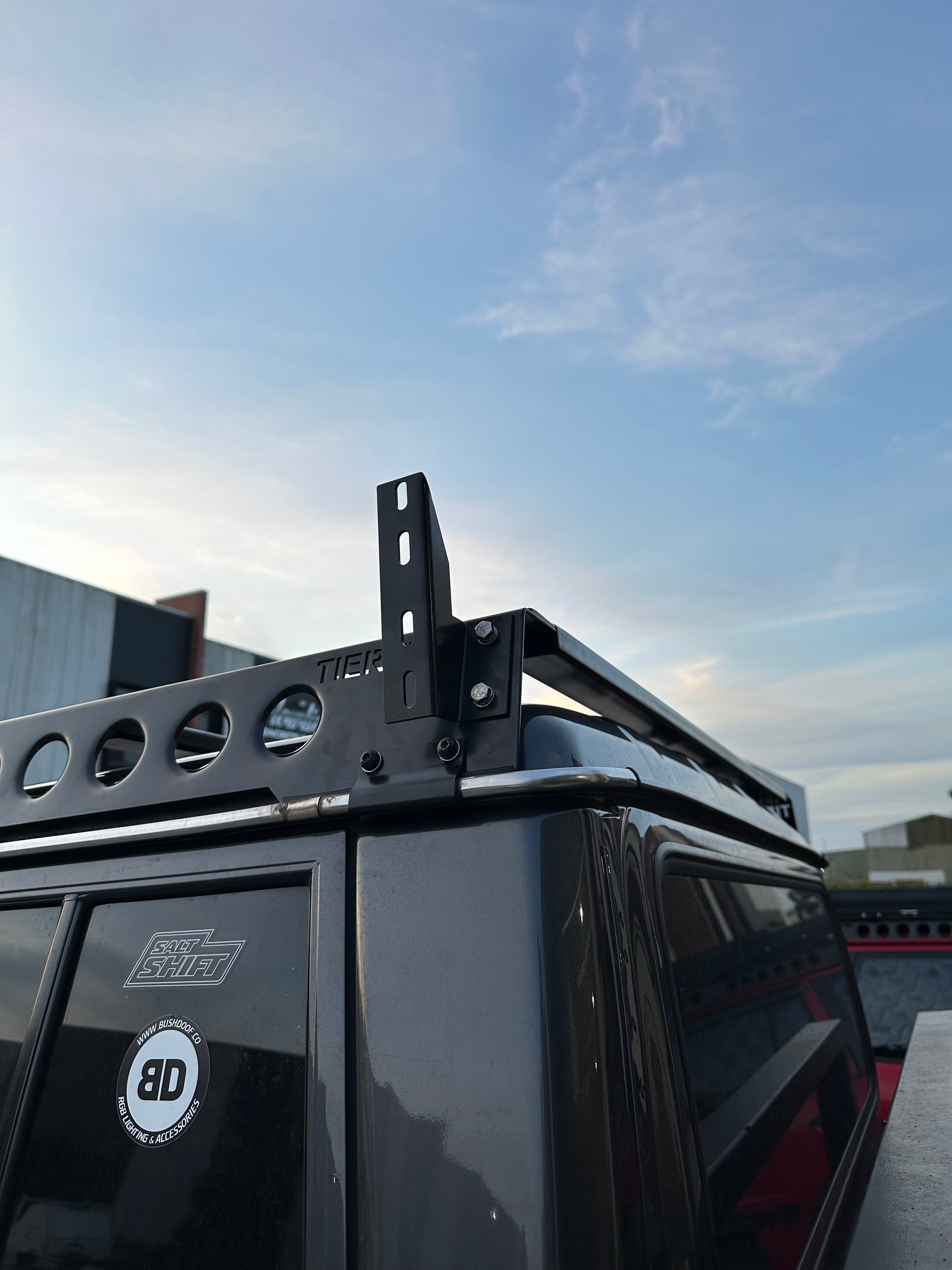 This is an image of a black Tier One Metalworks roof rack mounted on a dark grey 4x4 with Salt Shift and Bushdoof Co stickers on the rear window