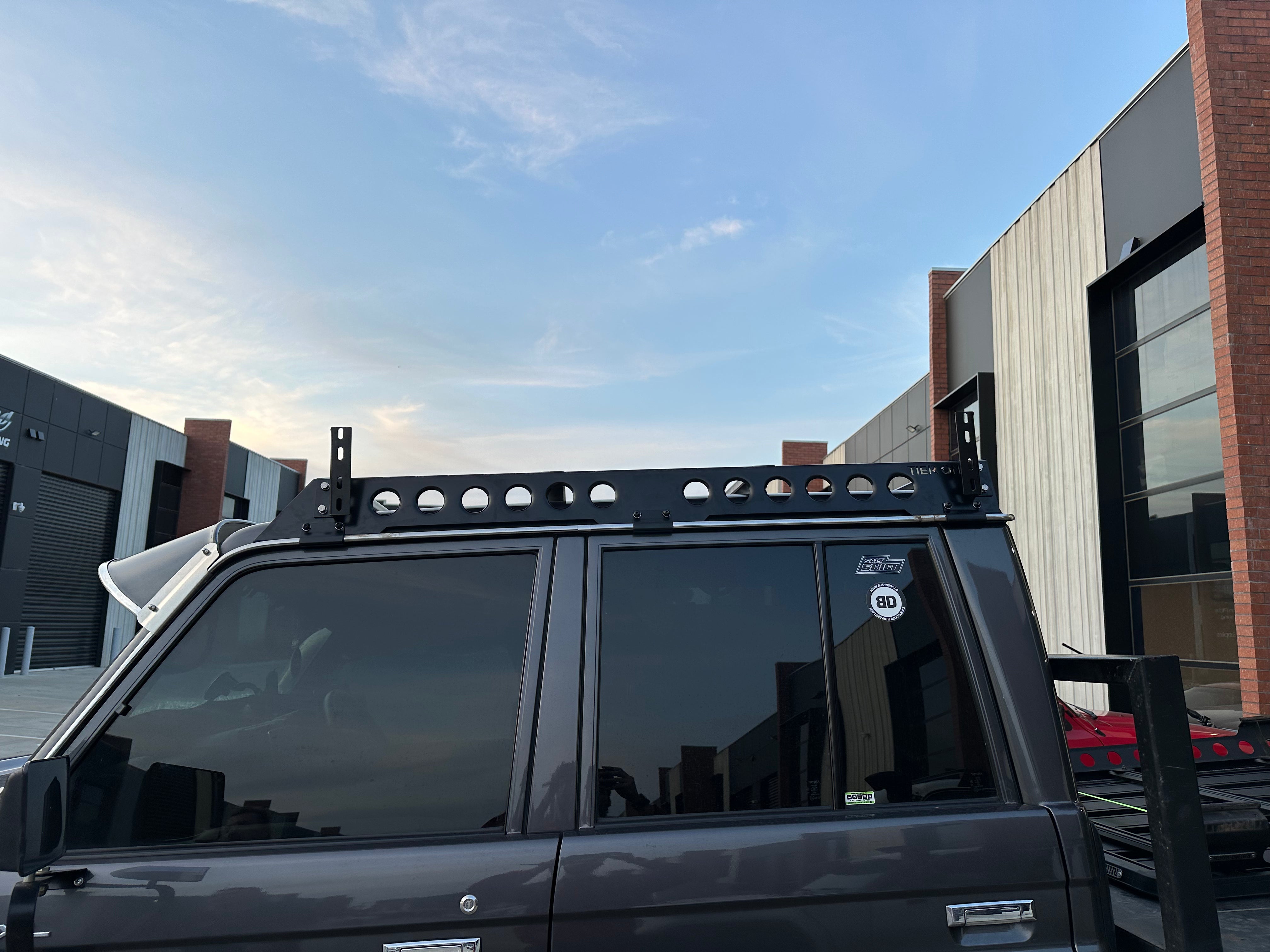 This is an image of a grey 4WD with Tier One Metalworks black roof racks installed, parked in an industrial area.