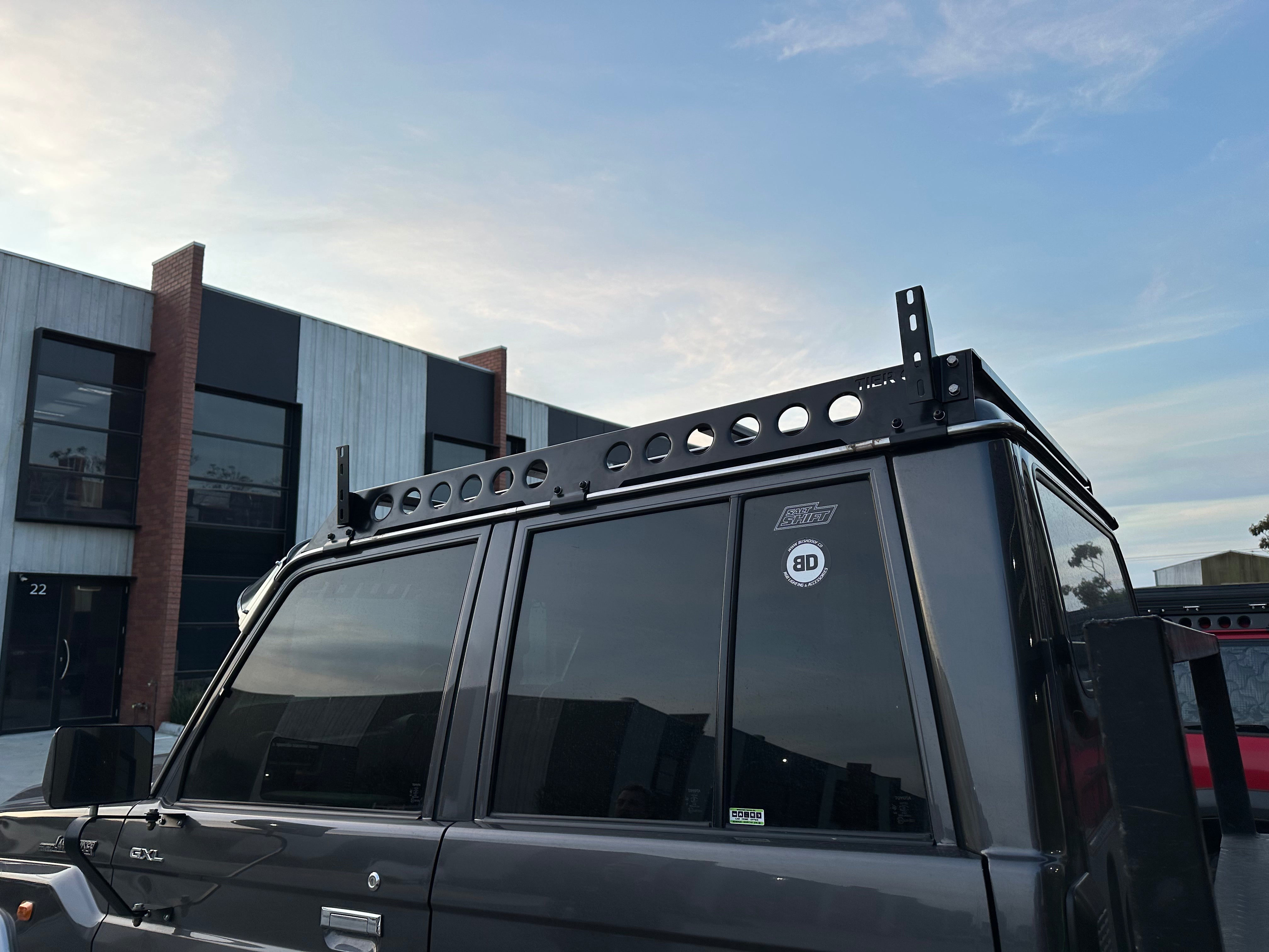 This is an image of a grey 4WD vehicle with Tier One Metalworks custom black roof racks and rails fitted, parked outside modern industrial units at dusk