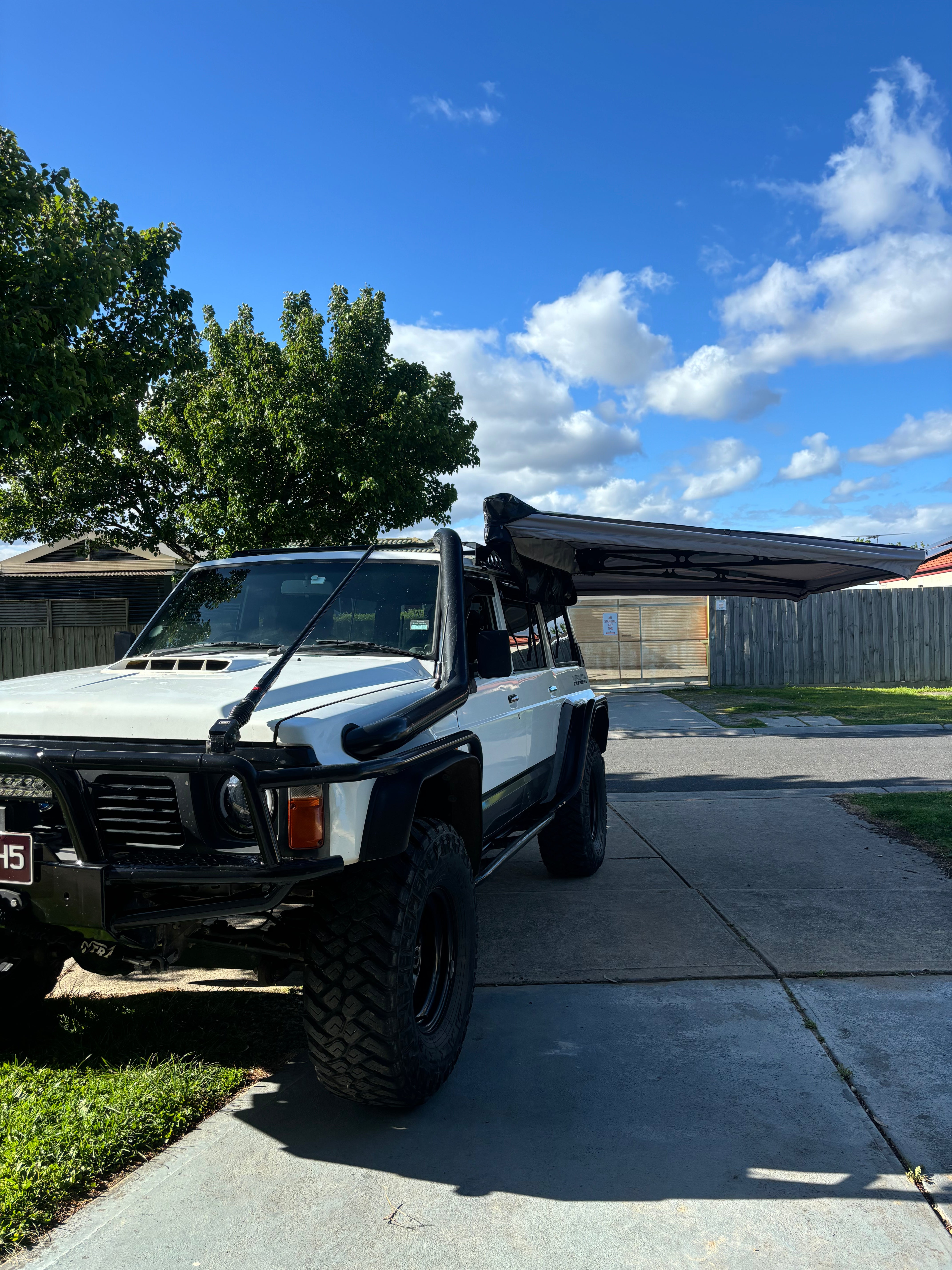 This is an image of a white 4WD with black snorkel and steel bull bar, fitted with a black Tier One Metalworks roof rack and extended awning under blue sky