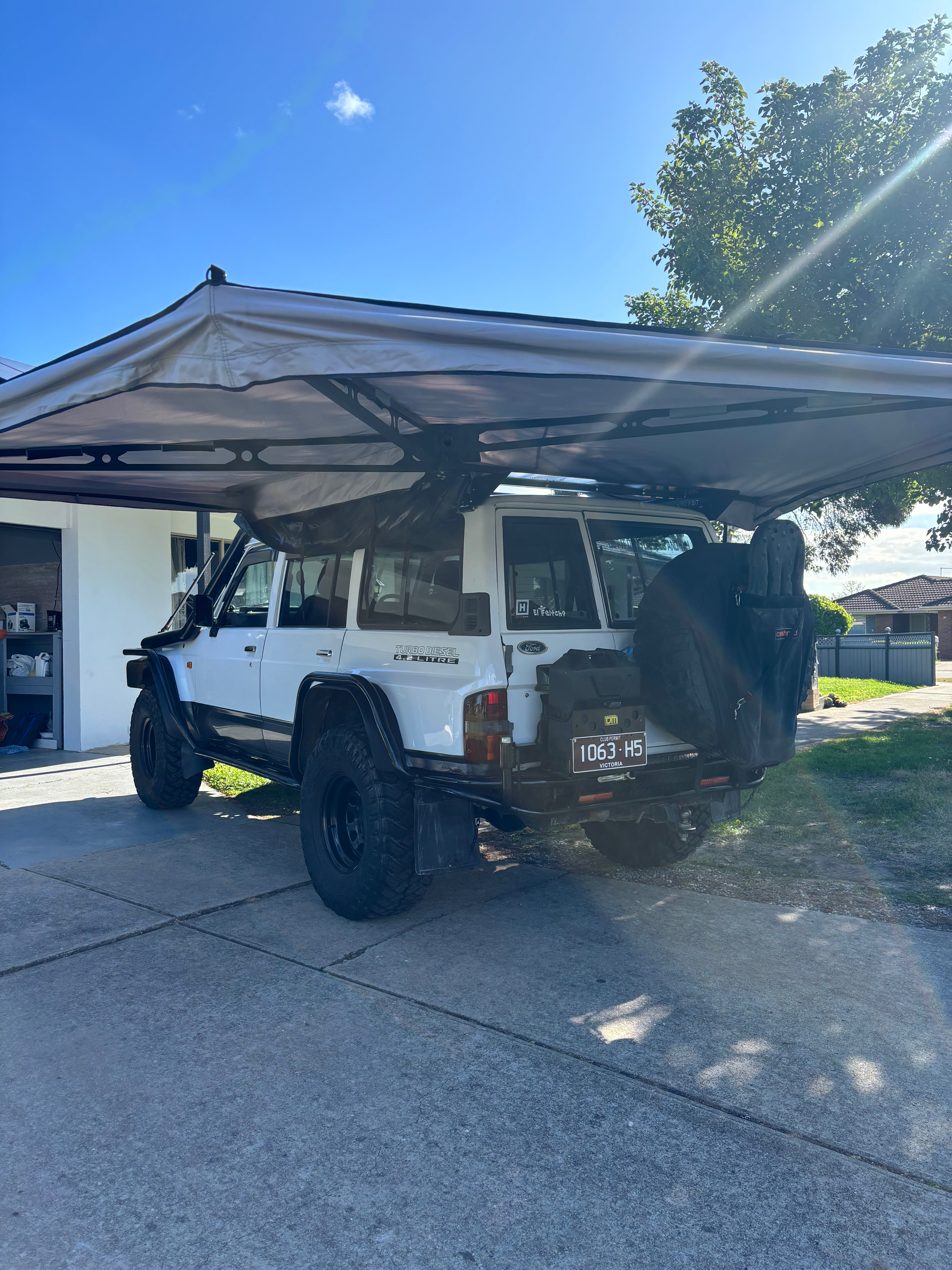This is an image of a white 4WD Ford Land Cruiser with an extended awning and Tier One Metalworks roof racks in a suburban driveway