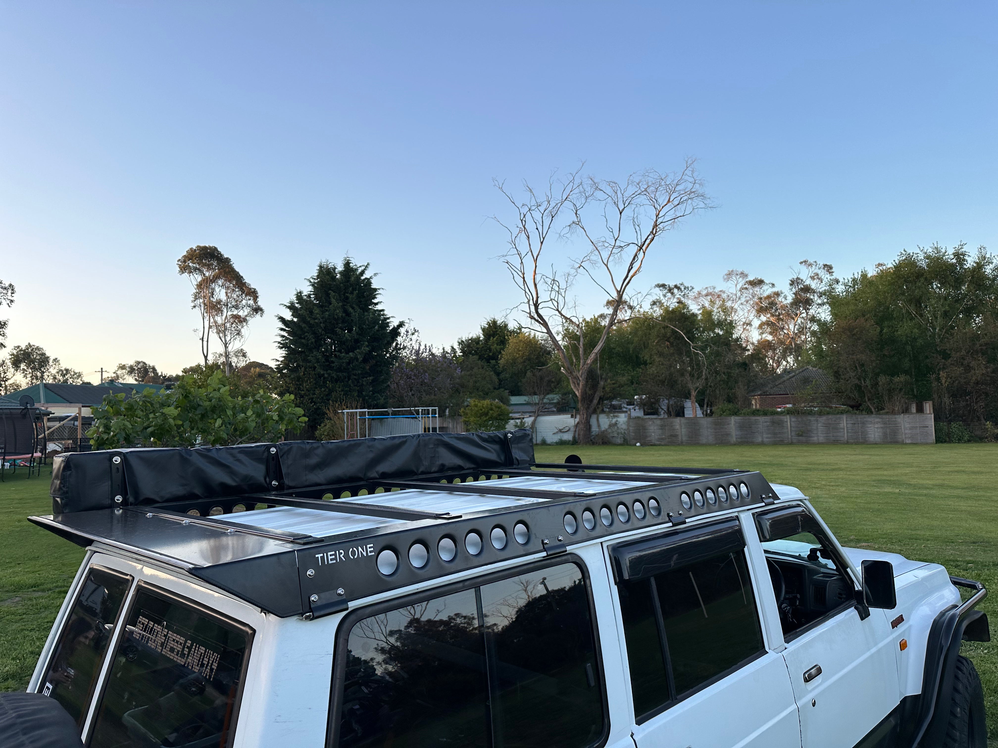 This is an image of a white 4WD with a Tier One Metalworks black roof rack and rails fitted, parked on grass.