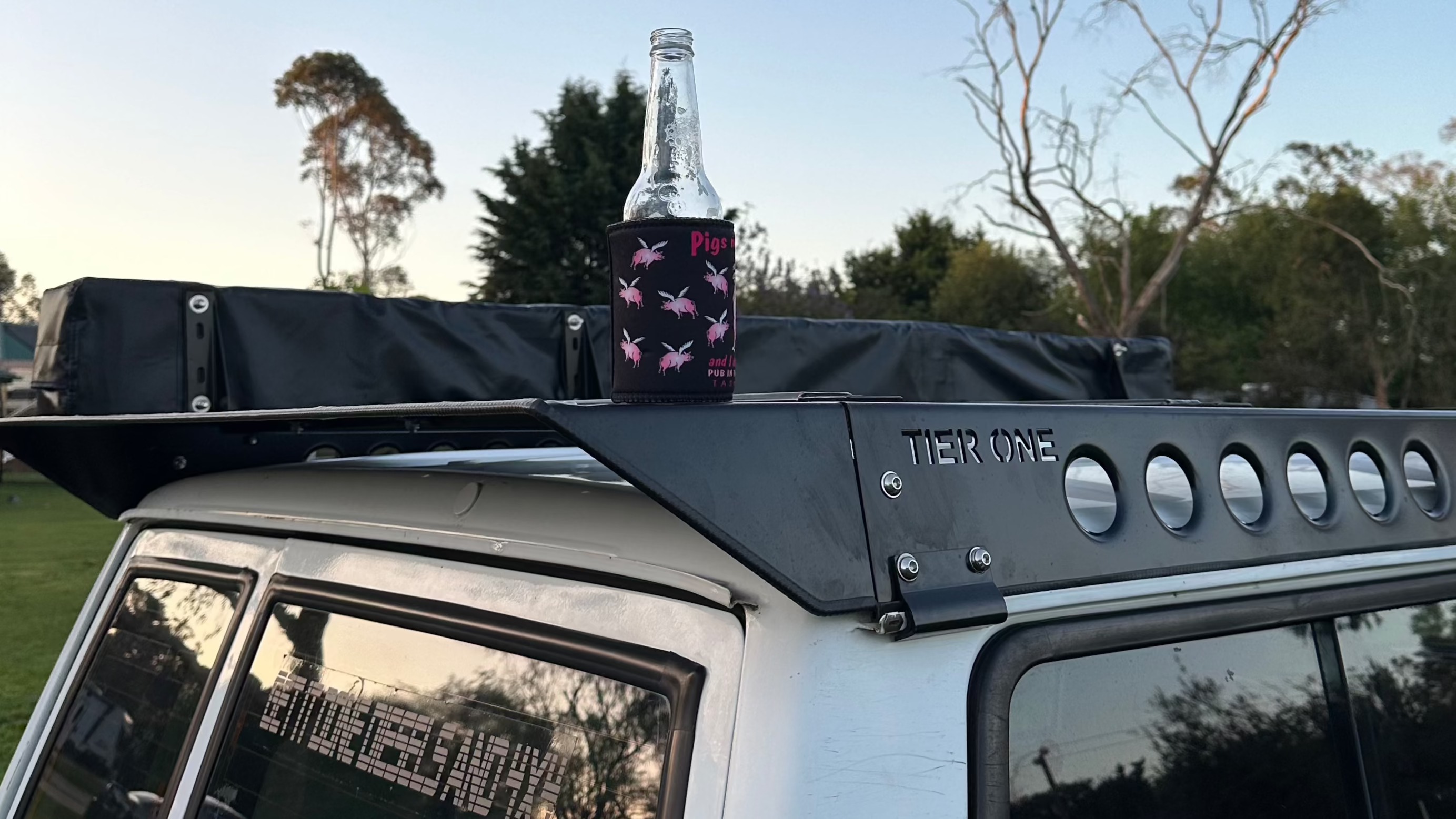 This is an image of a white 4x4 vehicle with a black Tier One Metalworks roof rack and a bottle with a koozie on top