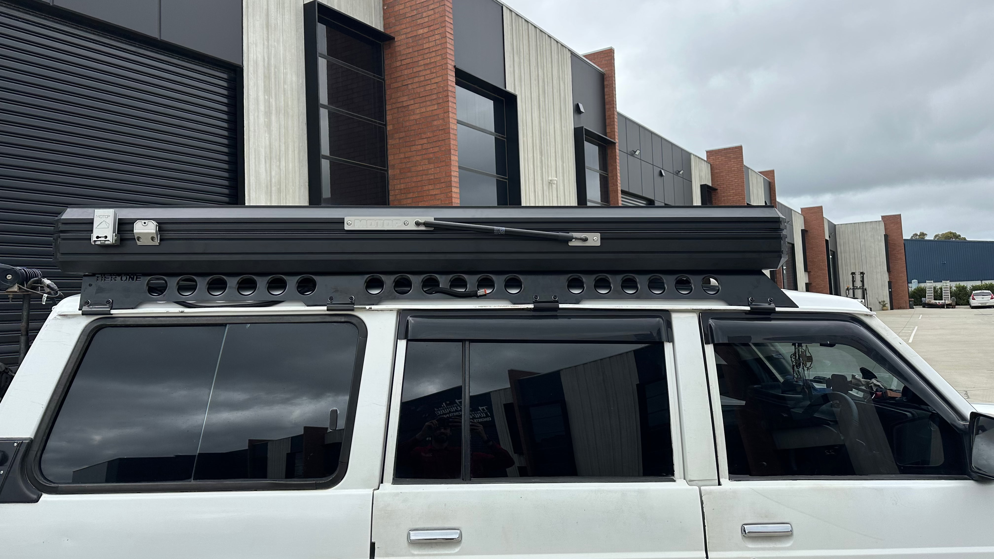 This is an image of a white 4WD with a black Tier One Metalworks roof rack and rooftop tent mounted, parked outside industrial units under a cloudy sky