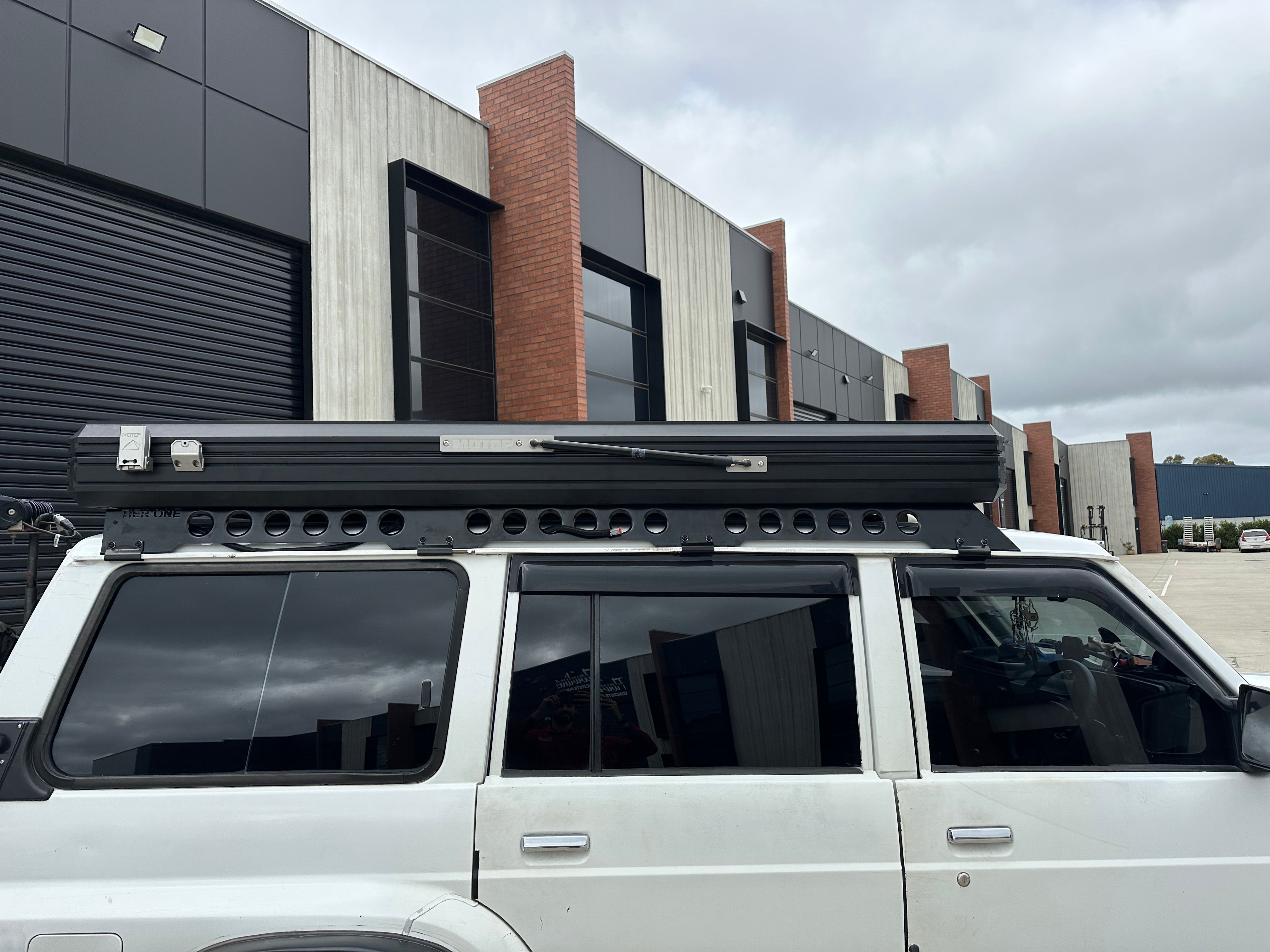 This is an image of a white 4WD with a black Tier One Metalworks roof rack and rooftop tent mounted, parked outside industrial units under a cloudy sky