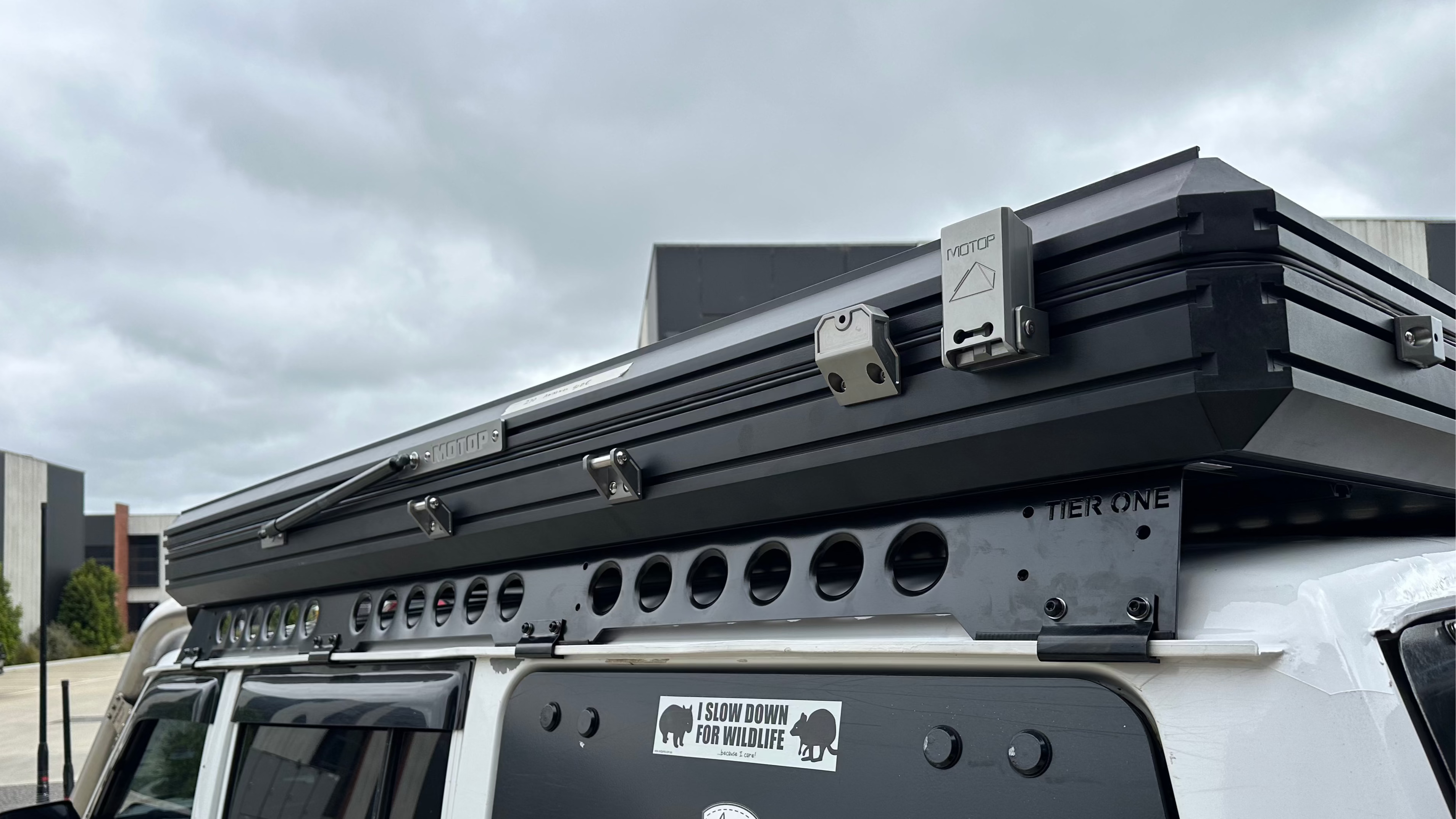 This is an image of a black Tier One Metalworks roof rack and Motop rooftop tent mounted on a white 4x4 vehicle under a cloudy sky