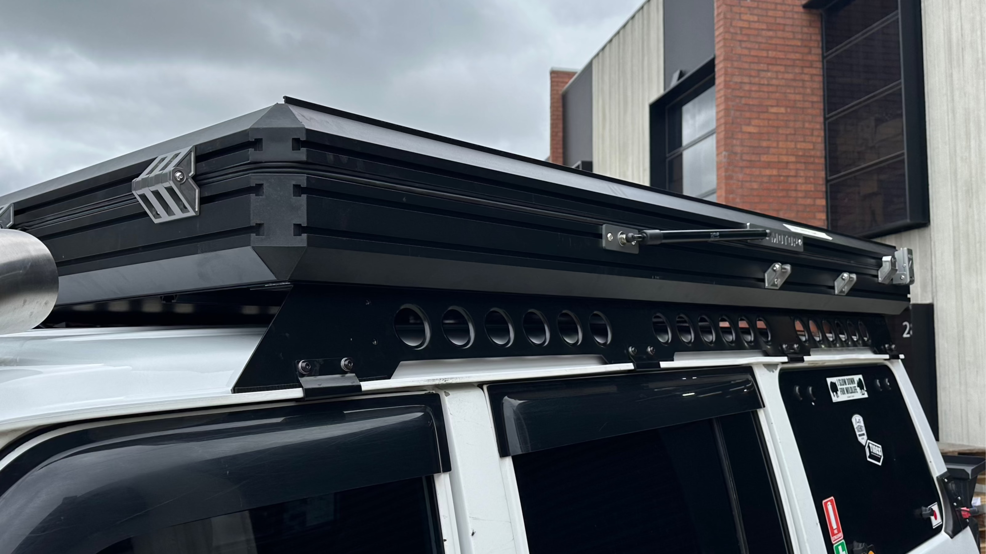 This is an image of a black Tier One Metalworks Roof Rack & Rails mounted on a white 4WD vehicle with tinted windows under overcast sky