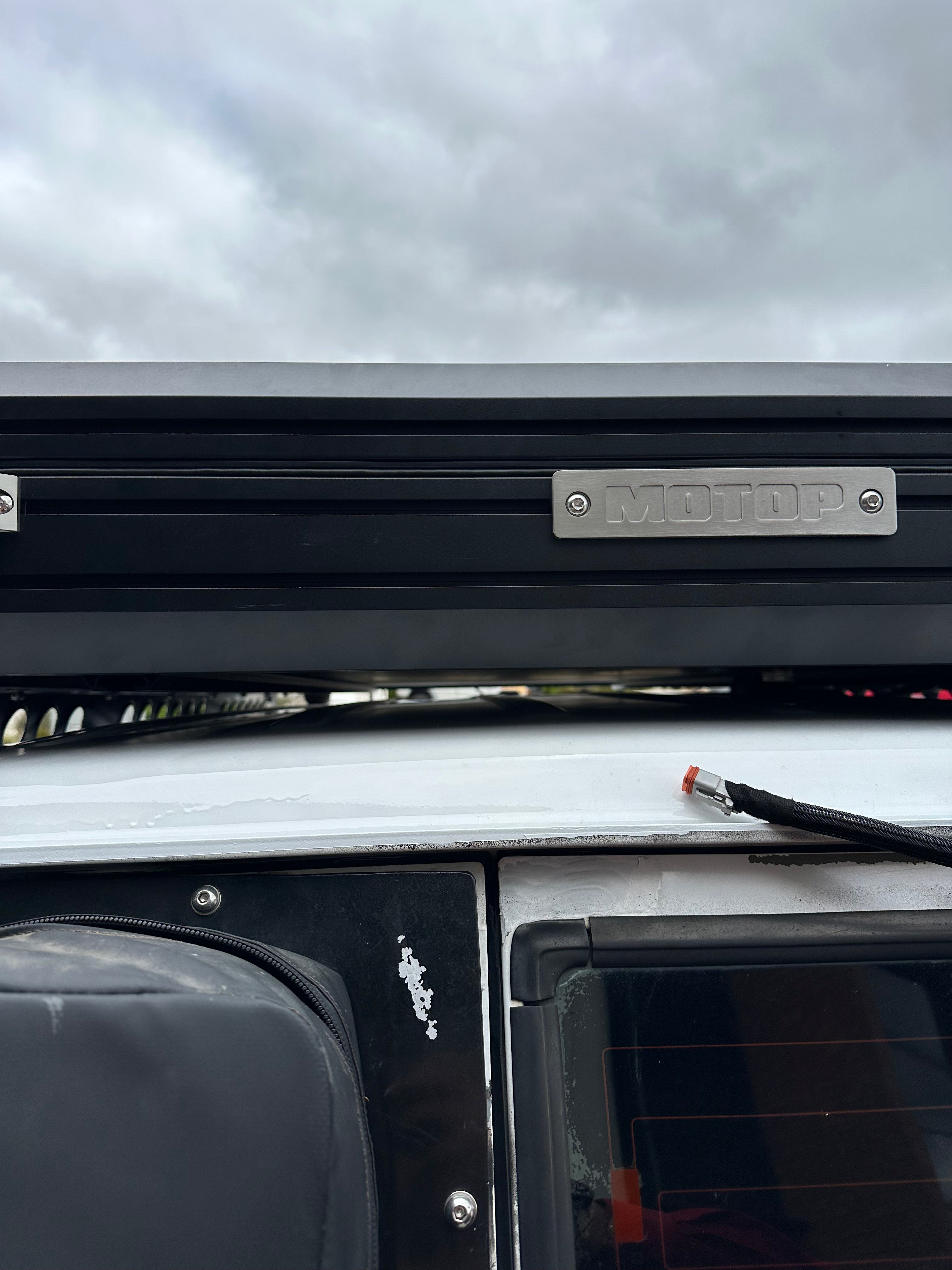 This is an image of a black Motop rooftop tent mounted on a white 4WD vehicle roof rack by Tier One Metalworks under an overcast sky