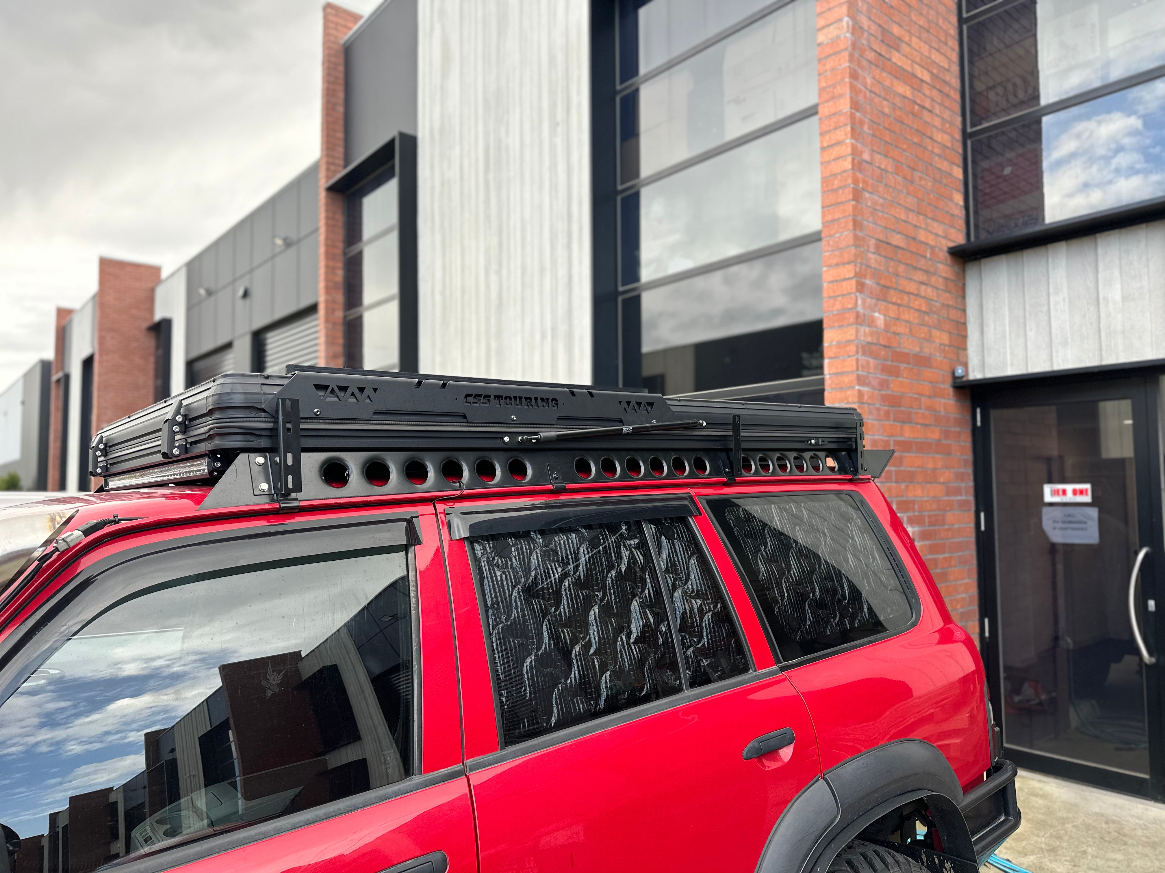 This is an image of a red 4WD fitted with a CSS Touring rooftop tent on a Tier One Metalworks roof rack and rails, outside an industrial building.