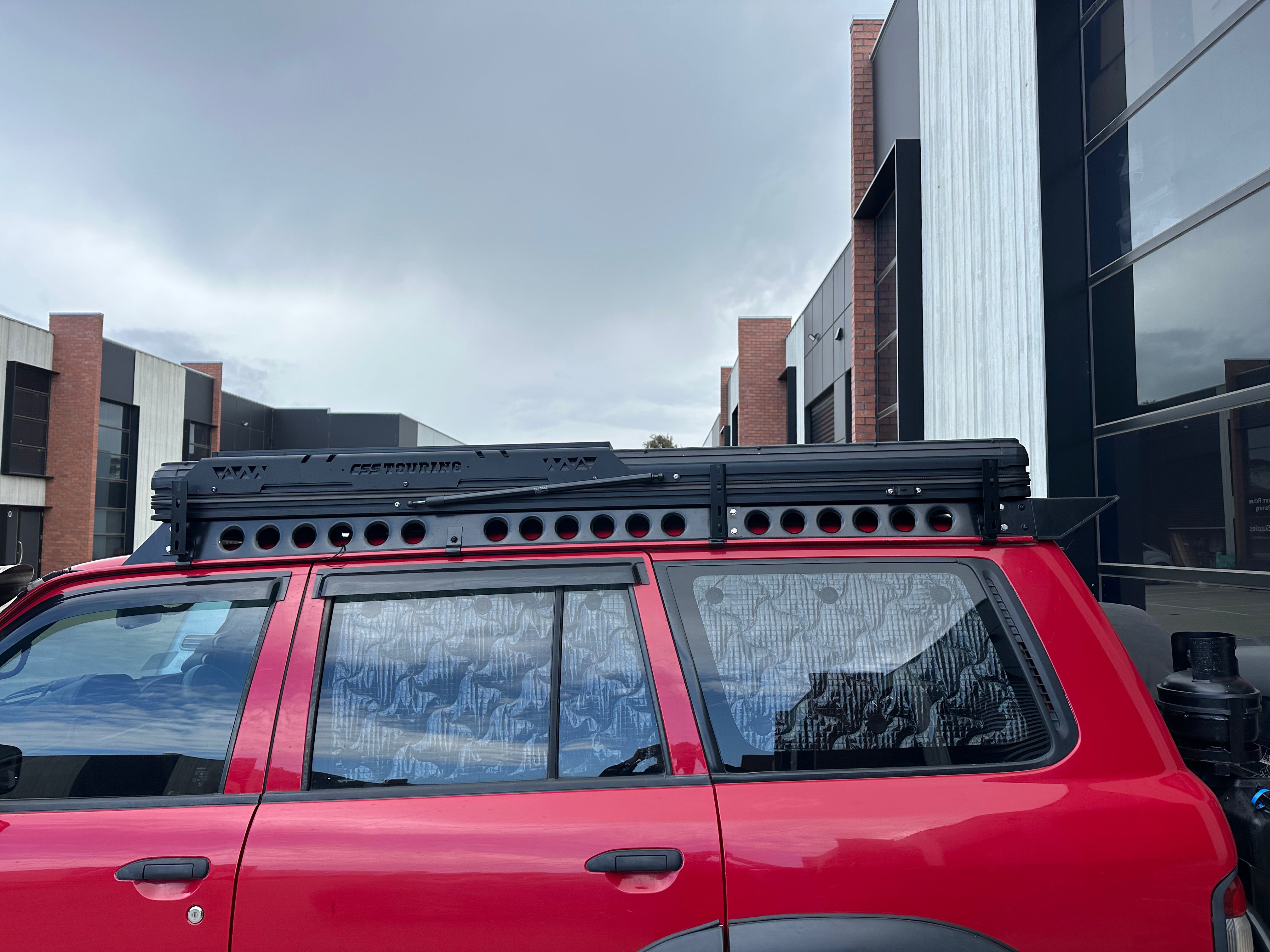 This is an image of a red 4WD fitted with a CSS Touring rooftop tent and Tier One Metalworks Roof Racks & Rails, parked in an industrial area.