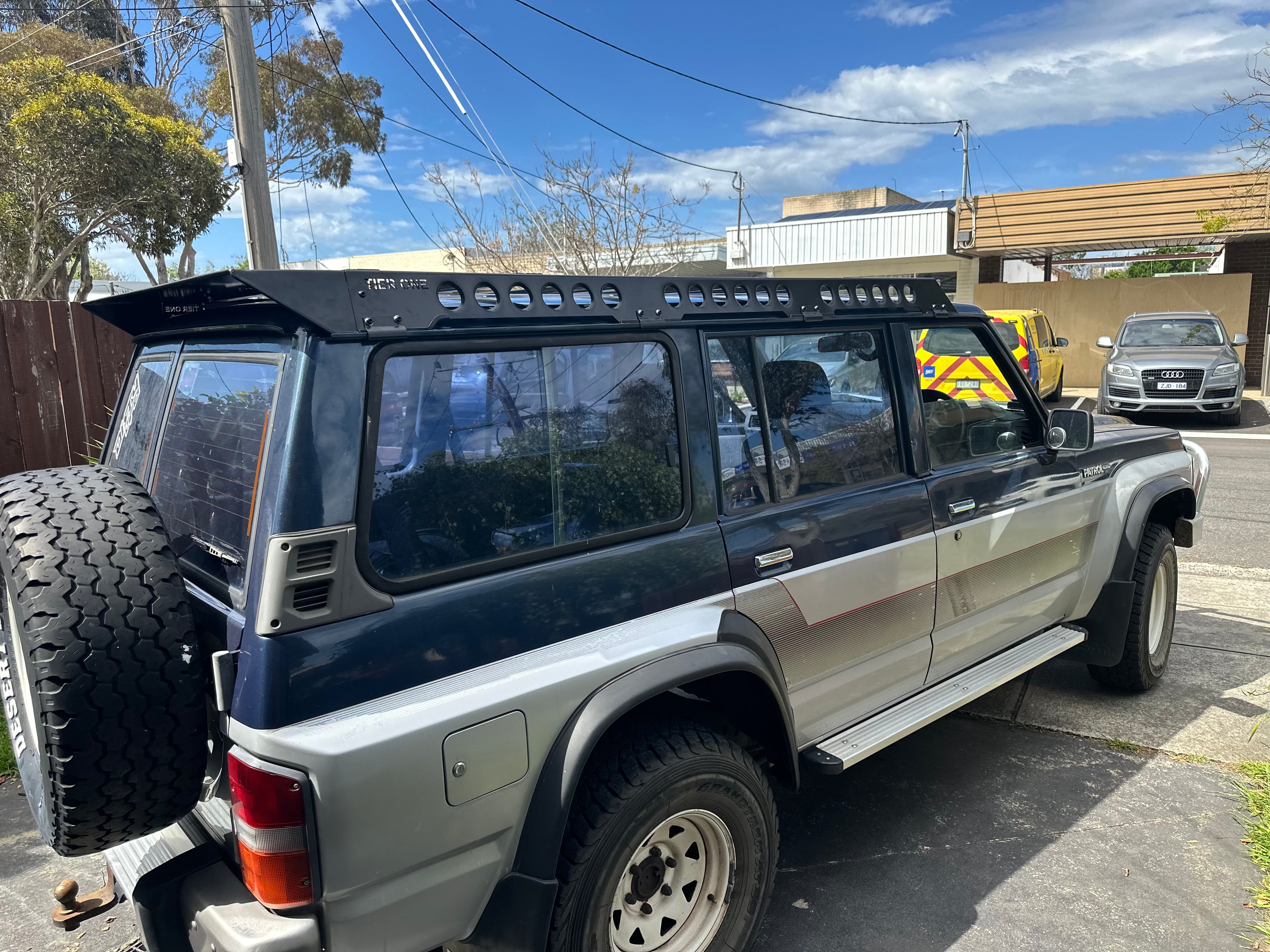This is an image of a grey and blue Nissan Patrol with a black Tier One Metalworks roof rack and rails parked in a suburban driveway