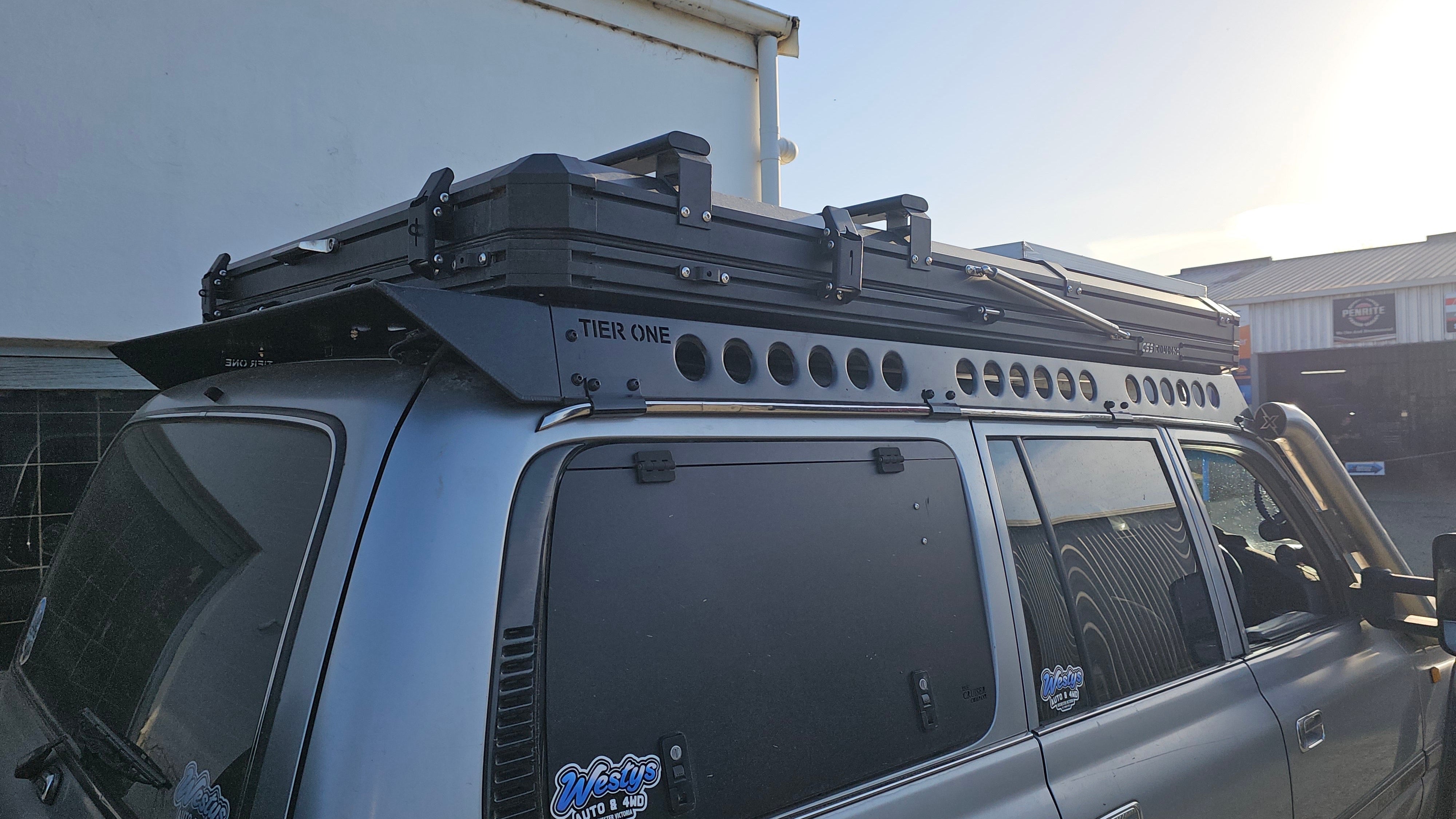 This is an image of a silver Toyota Land Cruiser 4WD with a Tier One Metalworks black roof rack and rails installed, parked outside in front of a building
