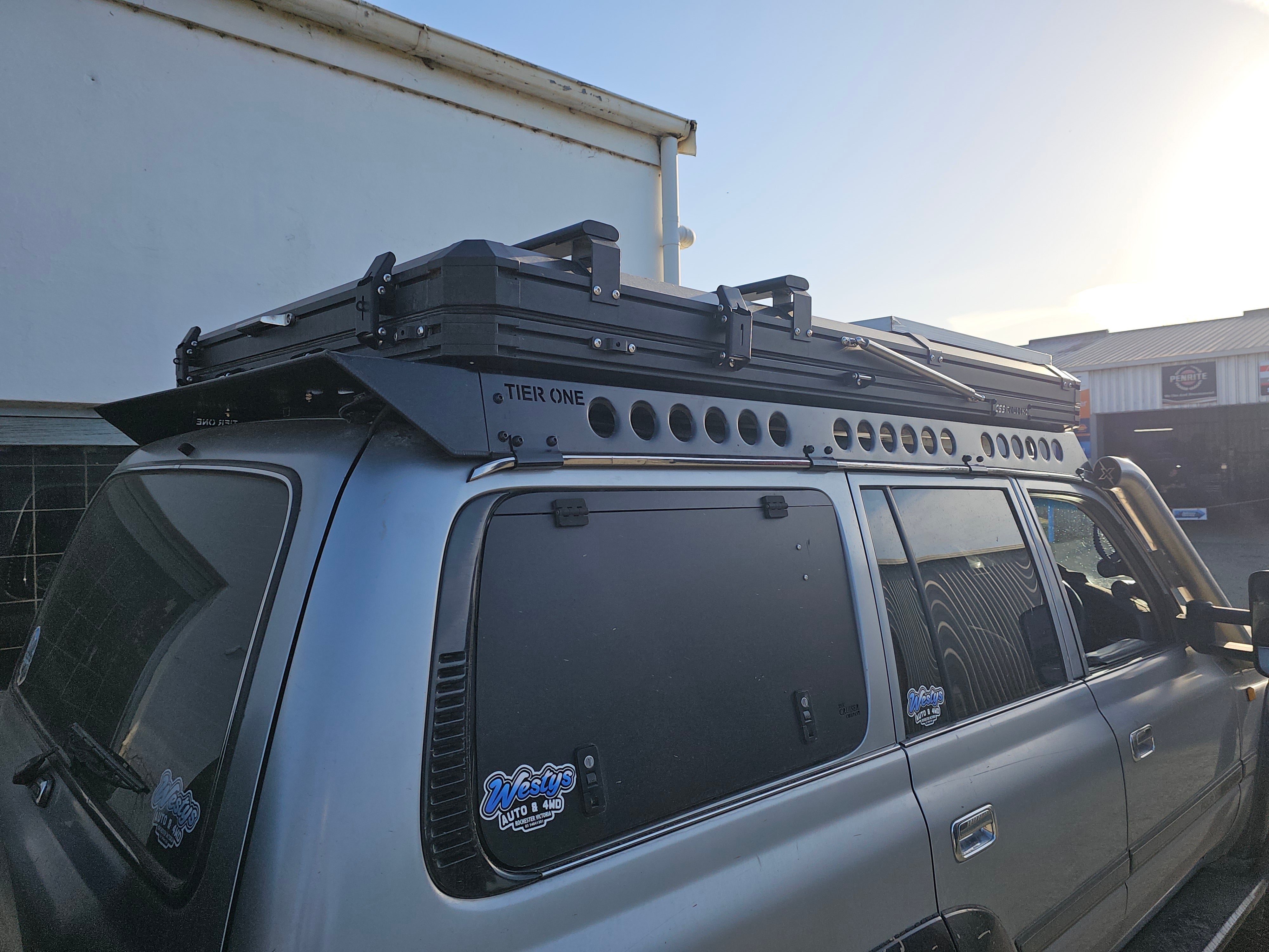 This is an image of a silver Toyota Land Cruiser 4WD with a Tier One Metalworks black roof rack and rails installed, parked outside in front of a building