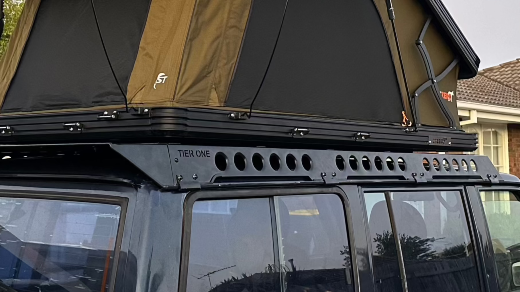 This is an image of a black Tier One Metalworks roof rack and rails mounted on a dark blue 4WD with a tan and black rooftop tent in a suburban setting