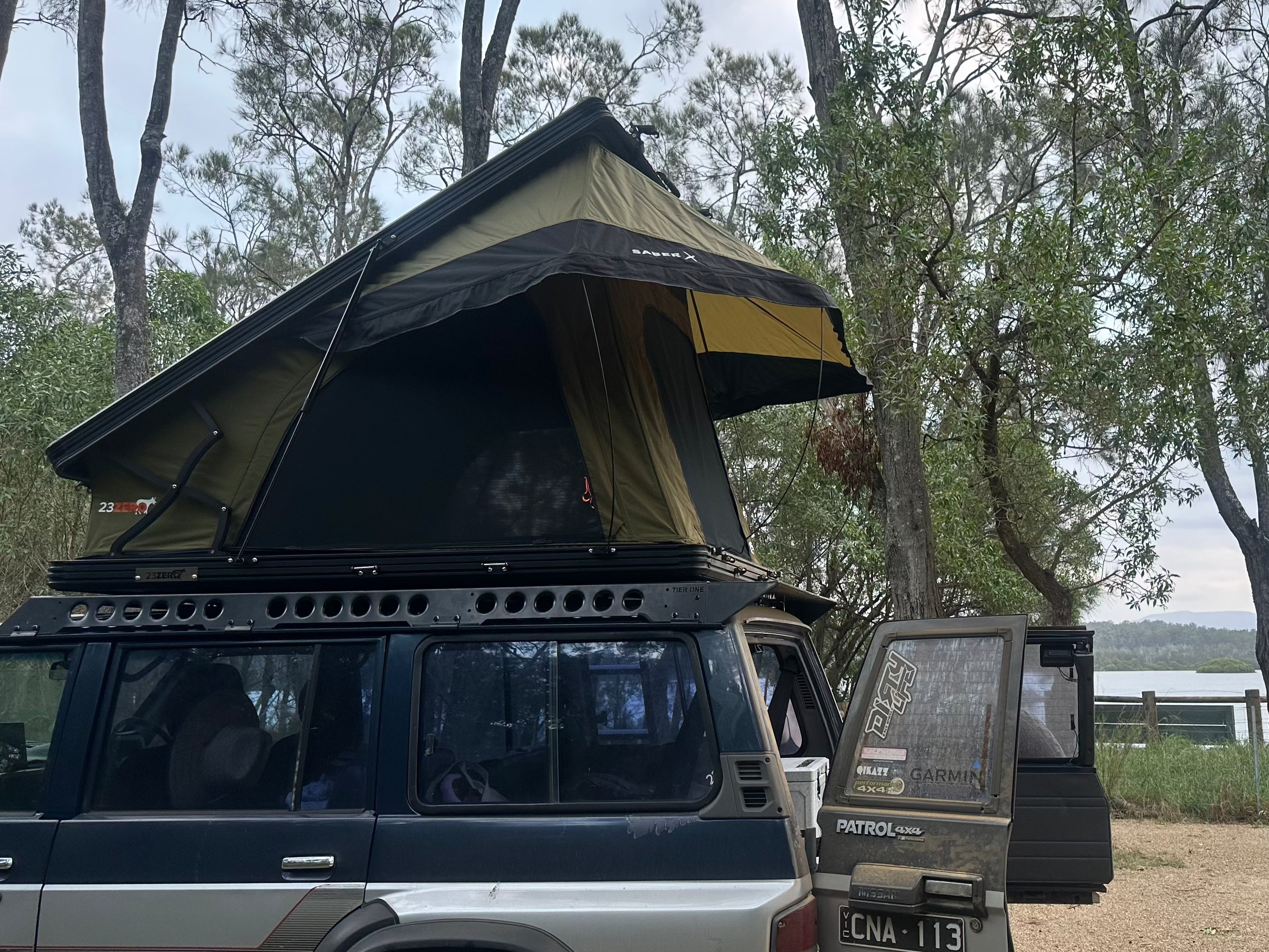 This is an image of a black and silver Nissan Patrol 4x4 with Tier One Metalworks roof racks and an open rooftop tent in a bushland setting