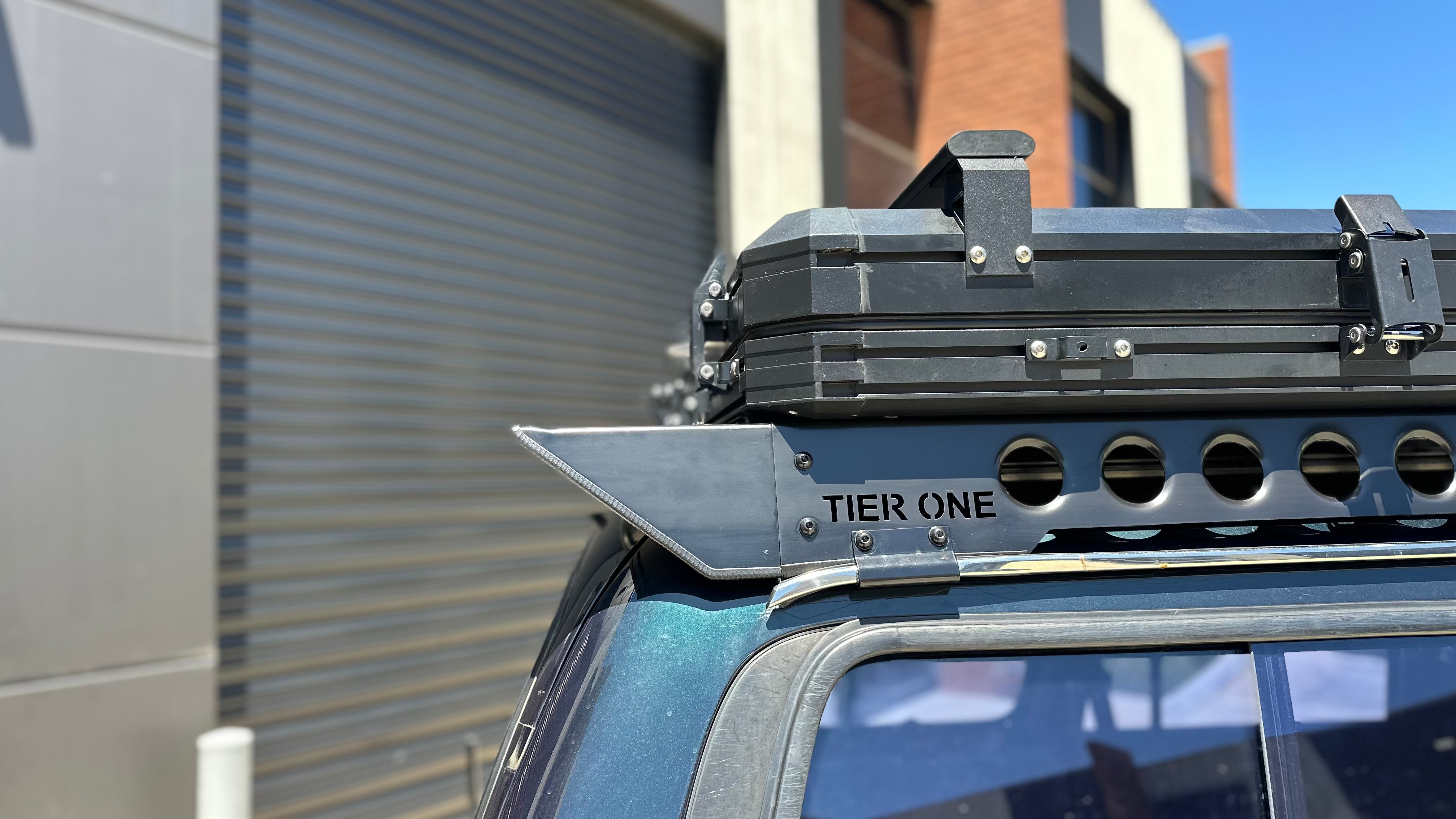 This is an image of a black Tier One Metalworks roof rack mounted on a dark blue 4x4 with a rooftop storage box, set against an industrial building background.