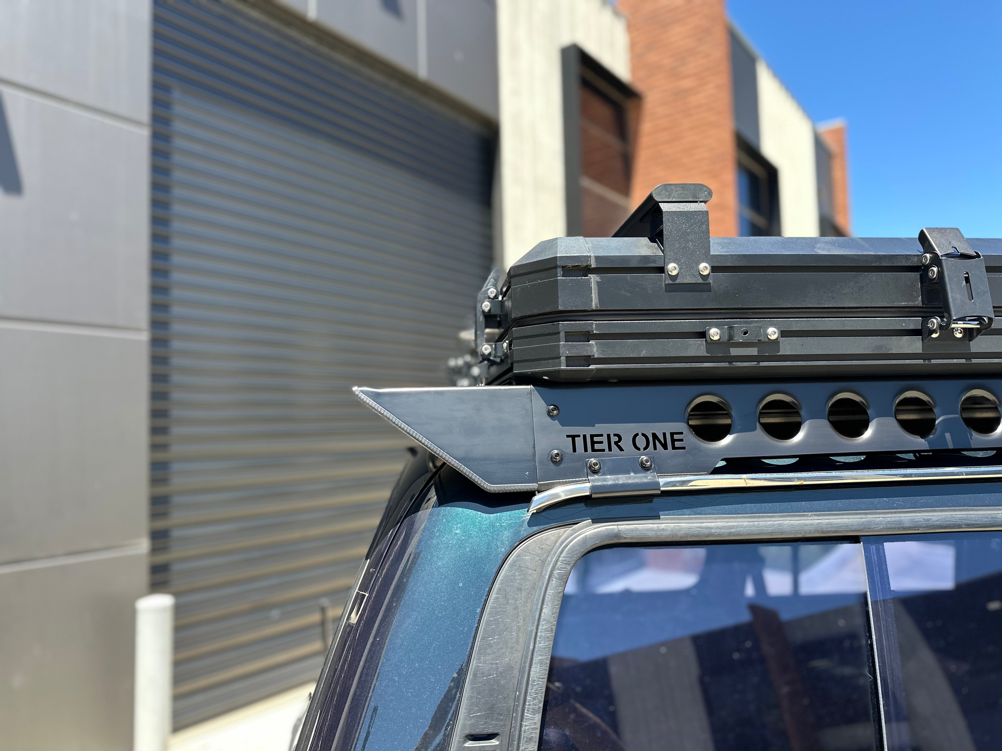 This is an image of a black Tier One Metalworks roof rack mounted on a dark blue 4x4 with a rooftop storage box, set against an industrial building background.