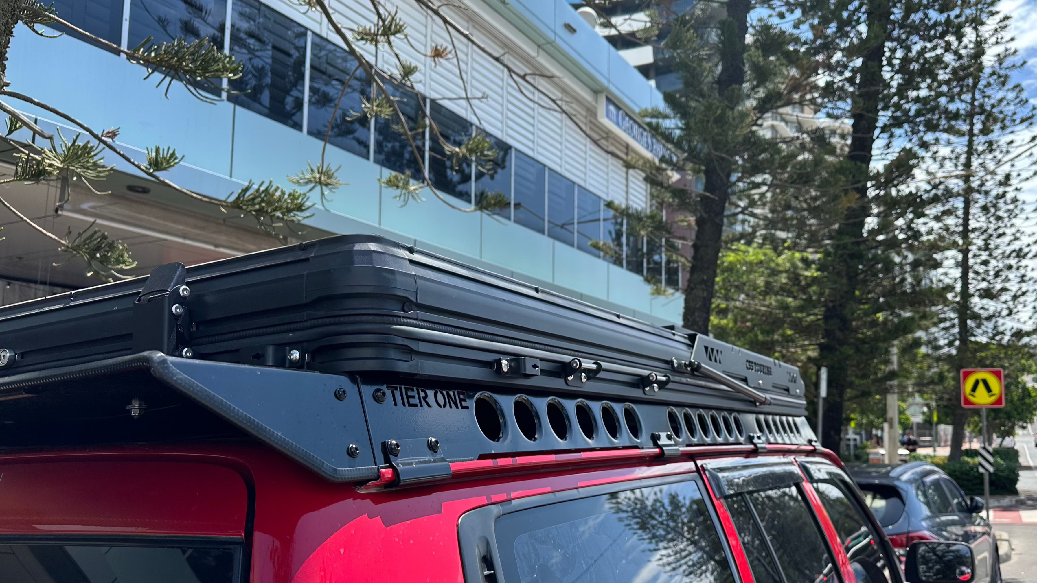 This is an image of a red 4x4 vehicle with a black Tier One Metalworks roof rack fitted on top in an urban setting
