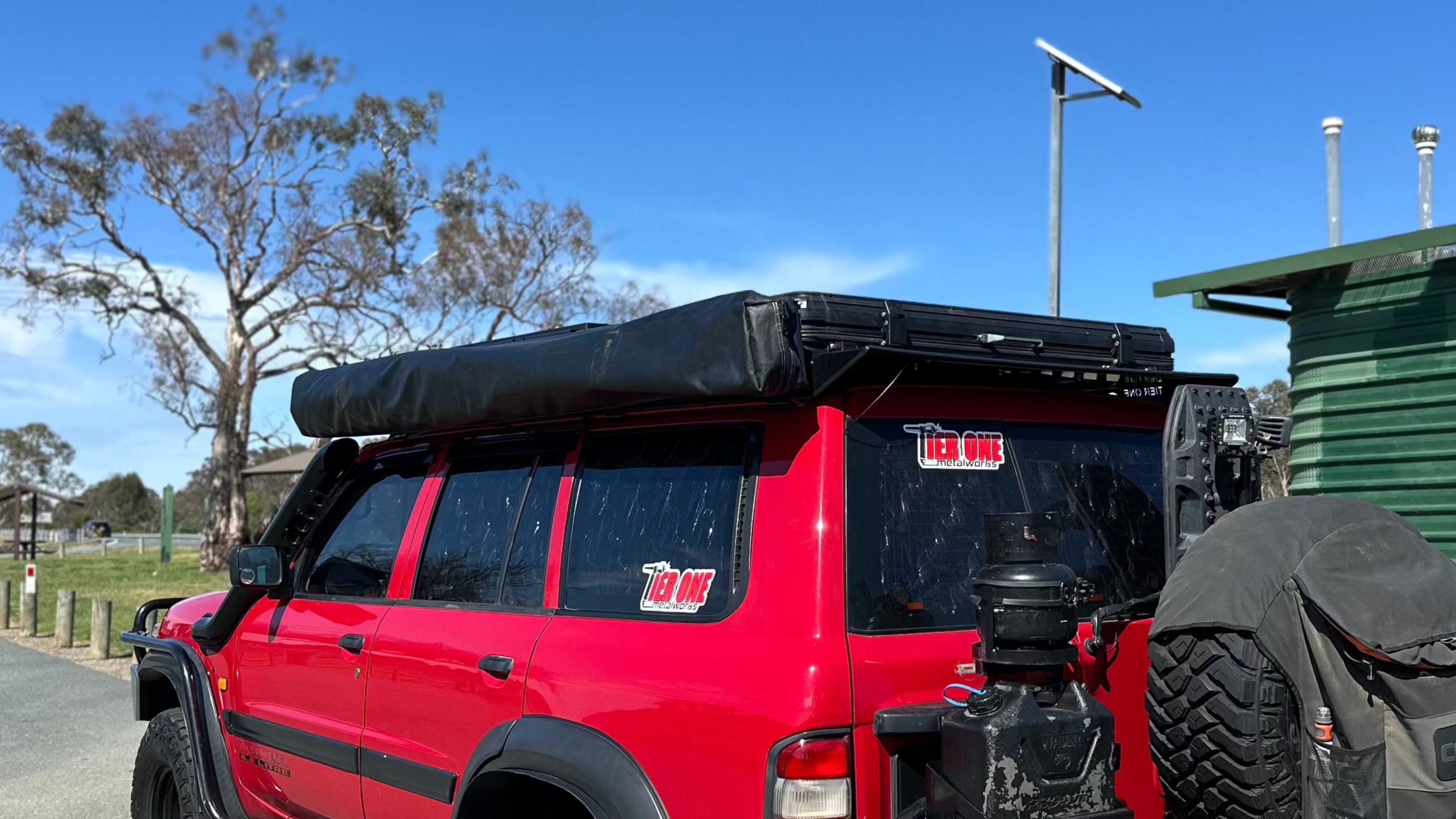 This is an image of a red 4x4 SUV with Tier One Metalworks roof racks and rails, fitted with a rooftop tent and rear spare tyre cover
