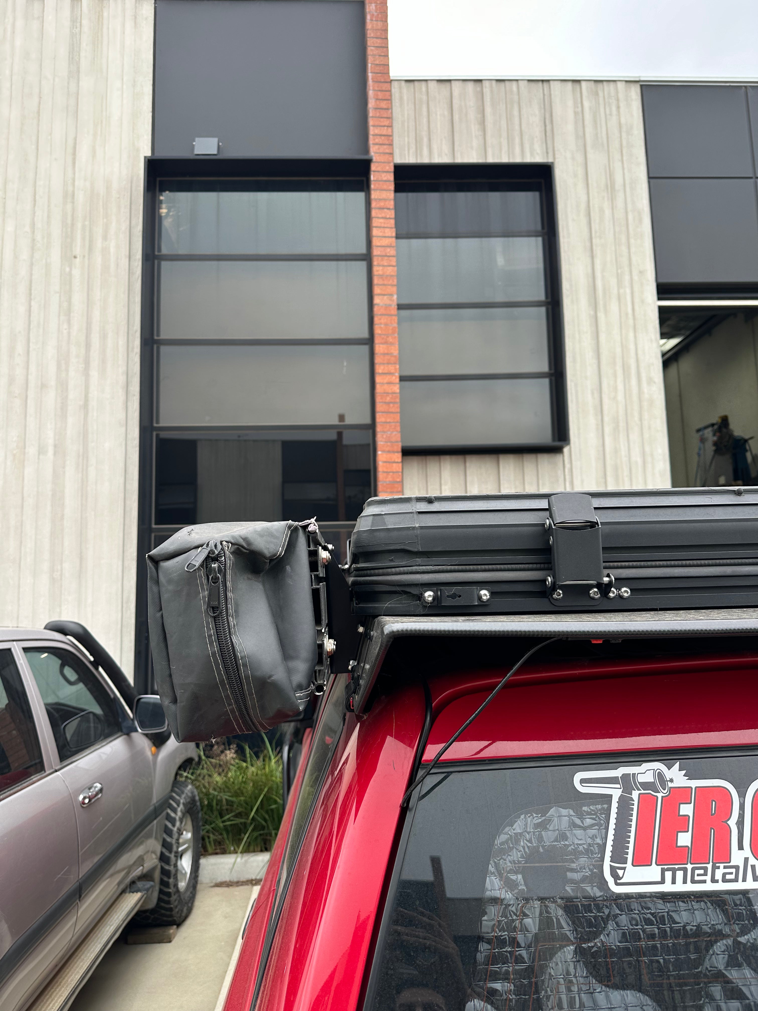 This is an image of a red 4x4 vehicle with a Tier One Metalworks Roof Rack fitted, showing a black accessory bag attached to the rack in an industrial setting.