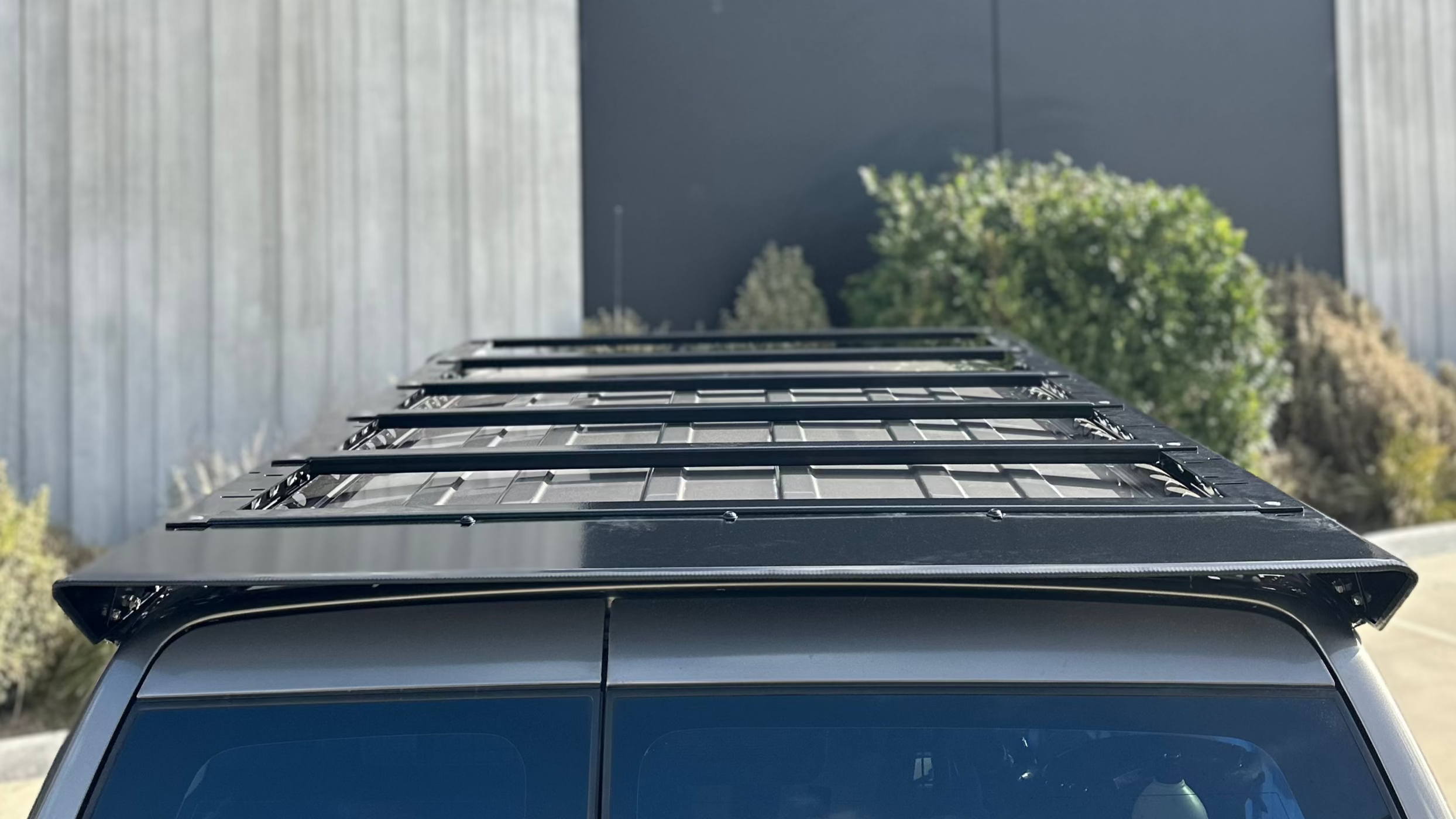 This is an image of a black Tier One Metalworks roof rack mounted on a silver 4x4 vehicle roof in an outdoor setting