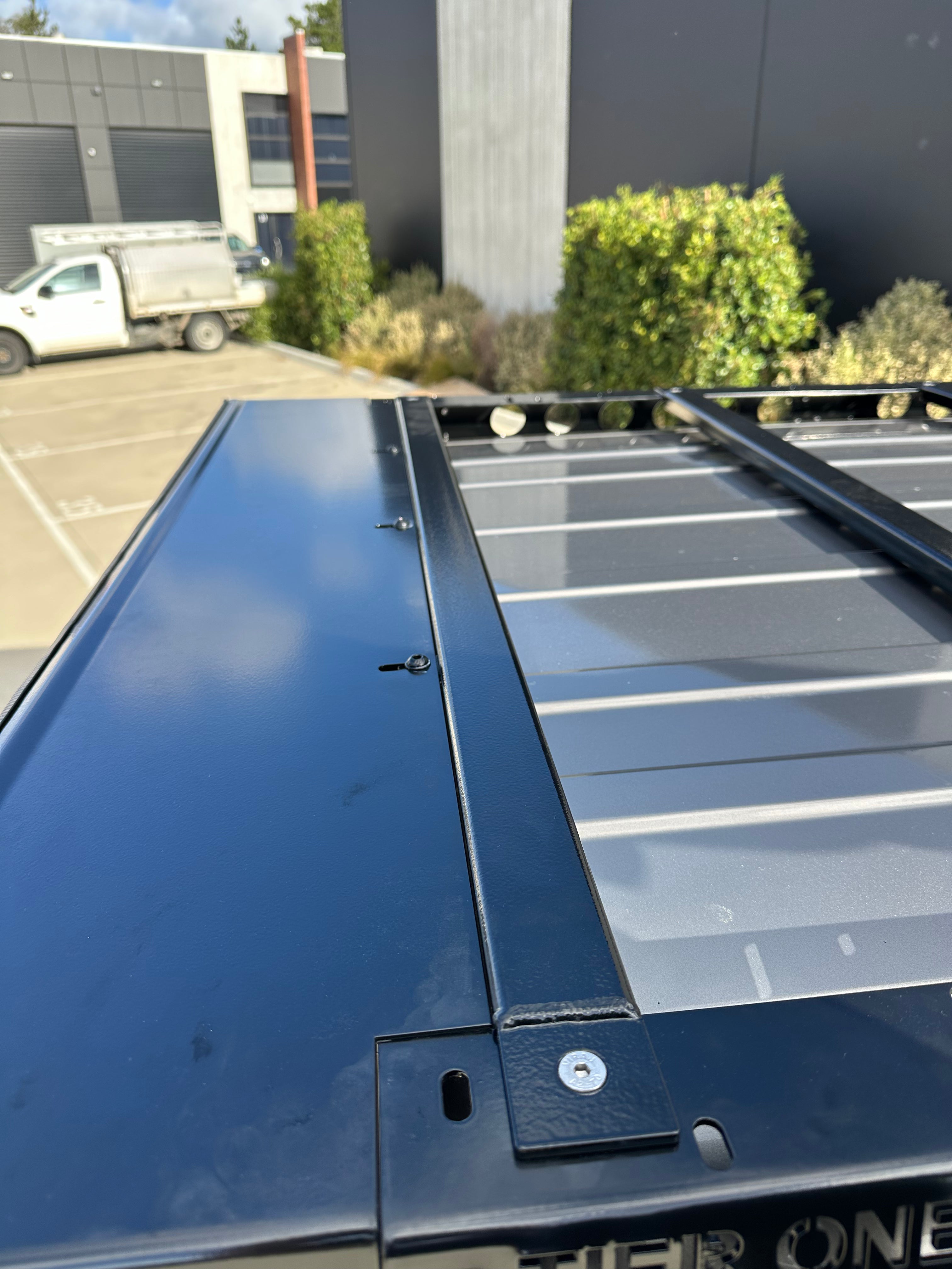 This is an image of a black Tier One Metalworks roof rack mounted on a vehicle roof with industrial buildings and greenery in the background