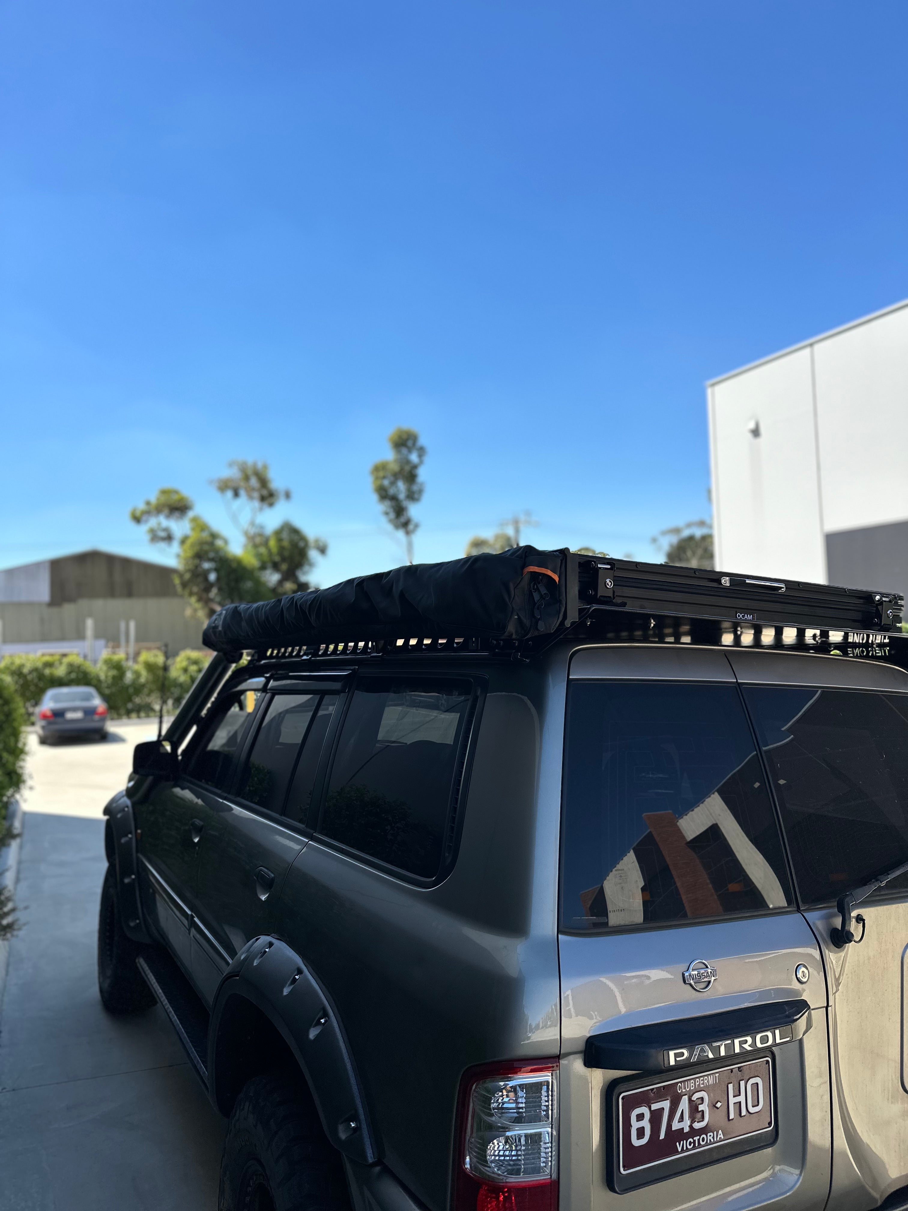 This is an image of a grey Nissan Patrol with Tier One Metalworks roof racks and rails parked outdoors under clear blue sky