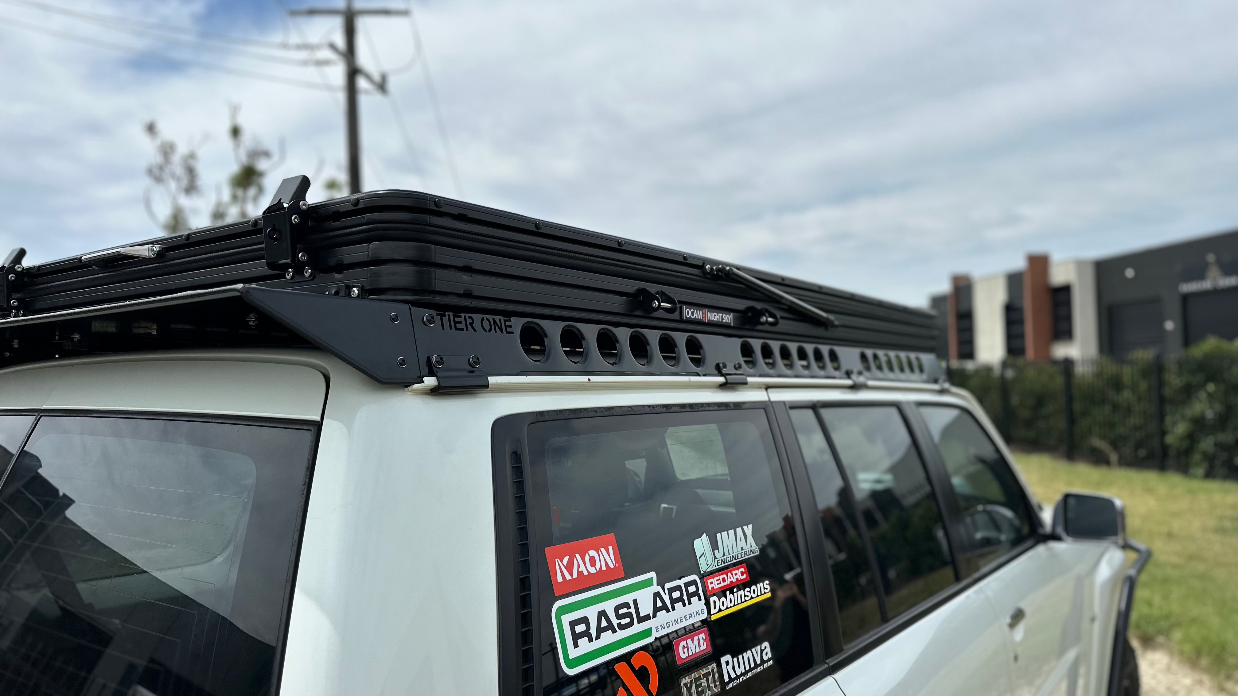 This is an image of a white 4x4 vehicle with Tier One Metalworks black roof racks and rails mounted, featuring multiple stickers on the rear side window including Raslarr and Dobinsons.