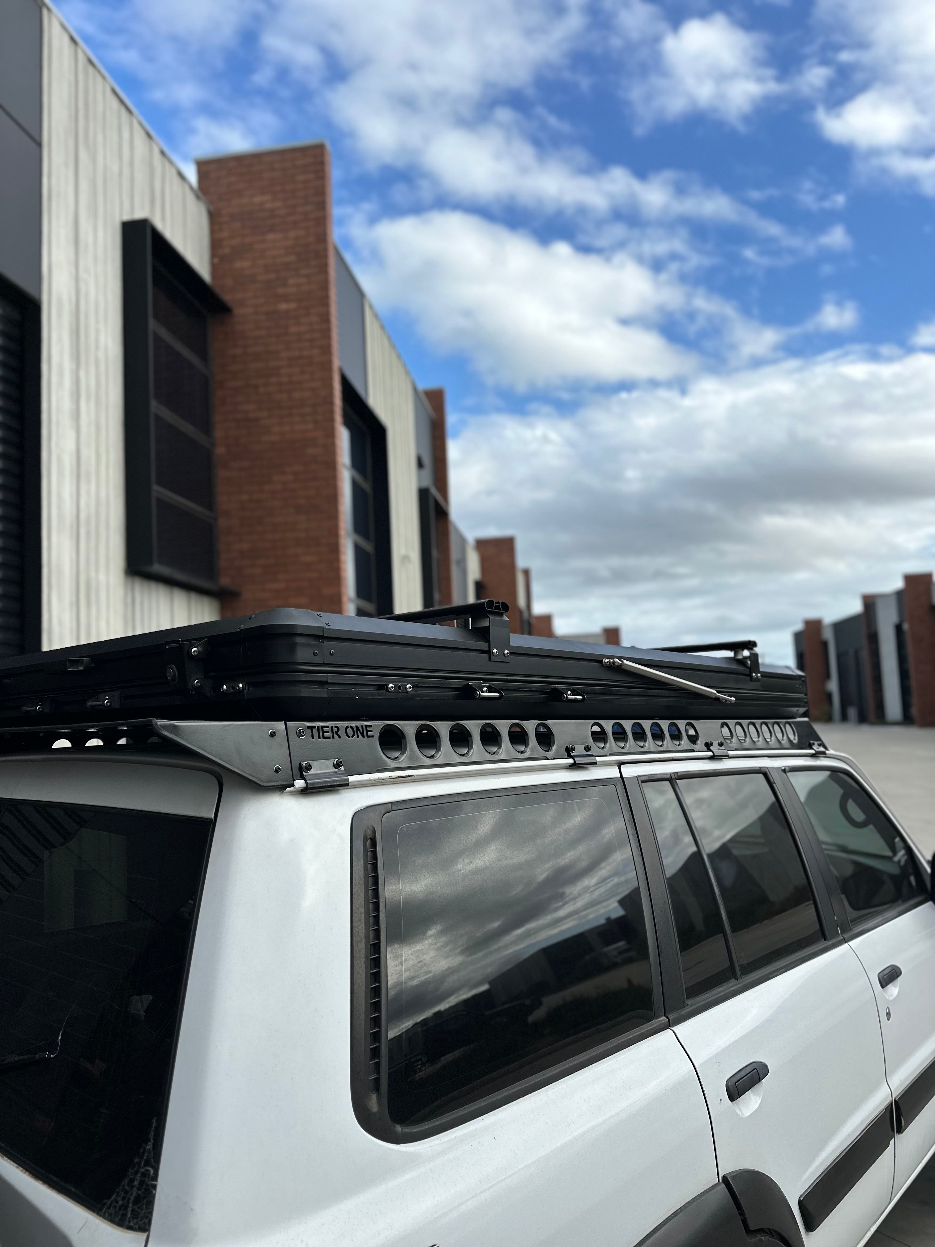 This is an image of a white 4x4 vehicle fitted with a black Tier One Metalworks roof rack and rails, parked near modern brick and concrete commercial buildings under a partly cloudy sky.