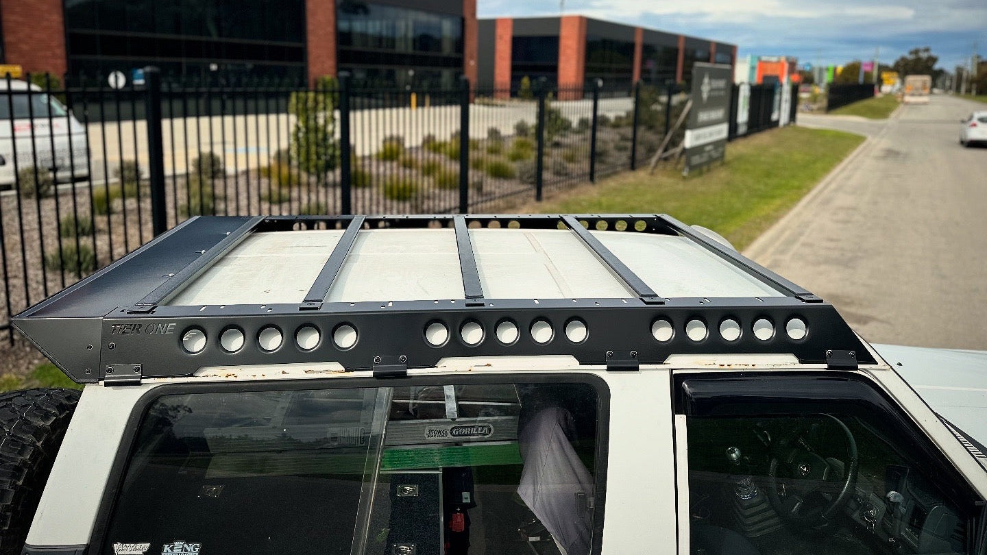 This is an image of a white 4x4 fitted with a black Tier One Metalworks Roof Rack on its roof, parked near an industrial area.