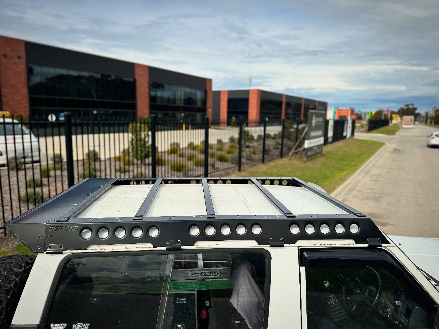 This is an image of a white 4x4 fitted with a black Tier One Metalworks Roof Rack on its roof, parked near an industrial area.