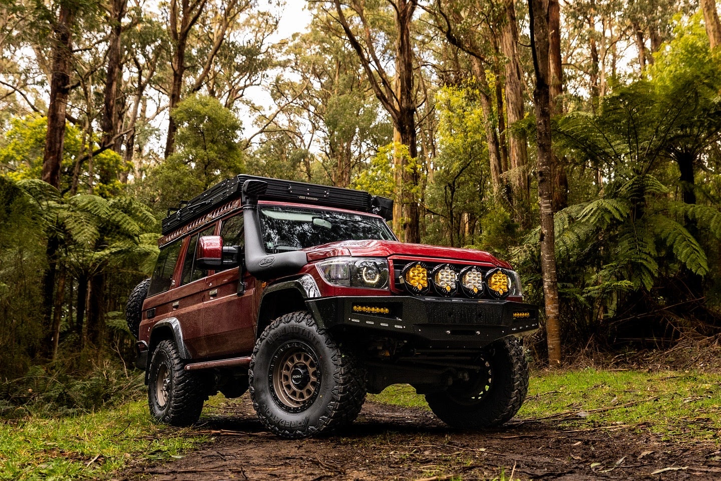 This is an image of a red Toyota Land Cruiser 4WD fitted with Tier One Metalworks roof racks and rails in a forest setting
