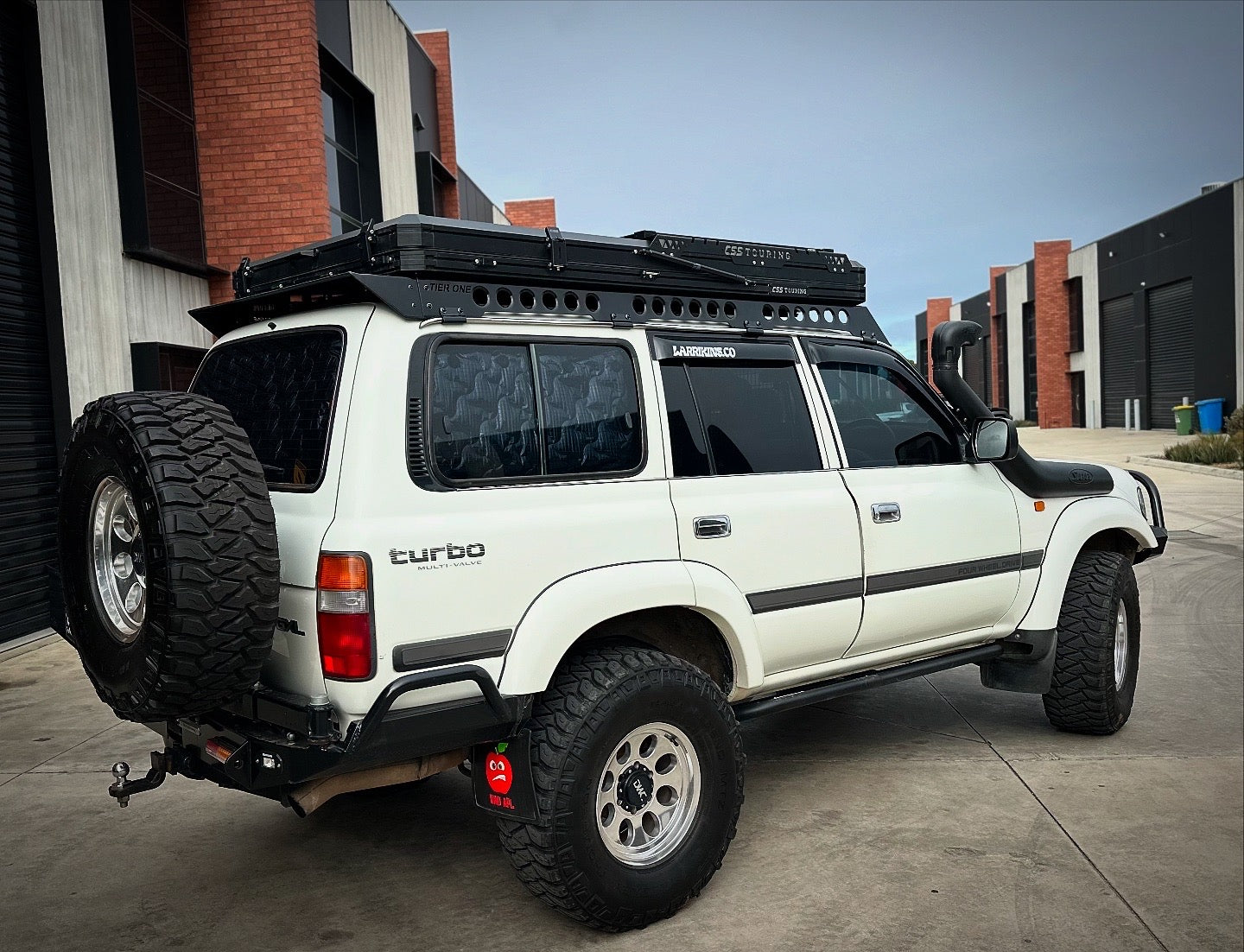 This is an image of a white Toyota Land Cruiser 80 Series with Tier One Metalworks roof racks and rails parked outside urban warehouses.